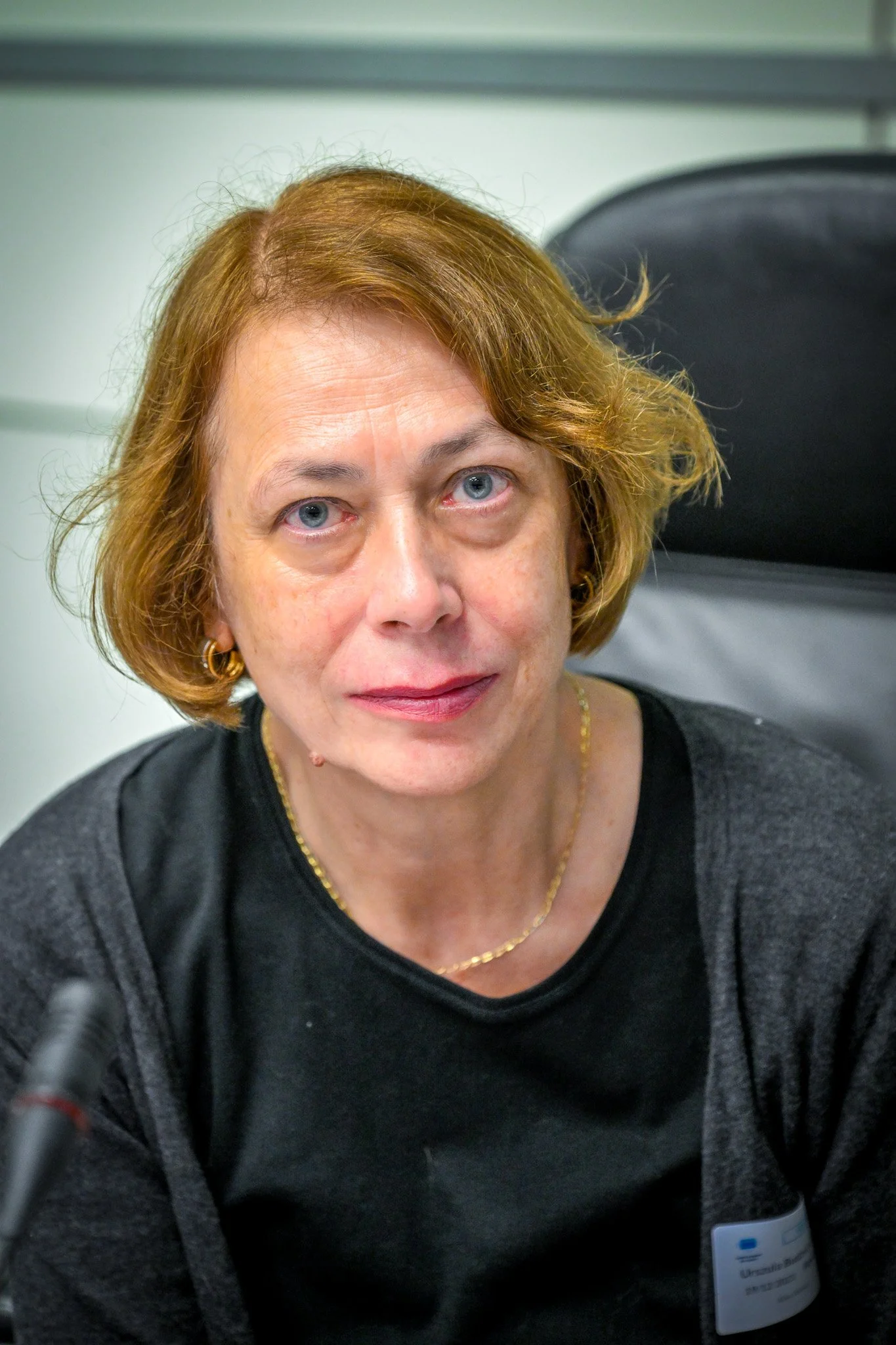 Femme avec des cheveux courts blonds roux, portant un collier et des boucles d'oreilles, assise sur une chaise noire.
