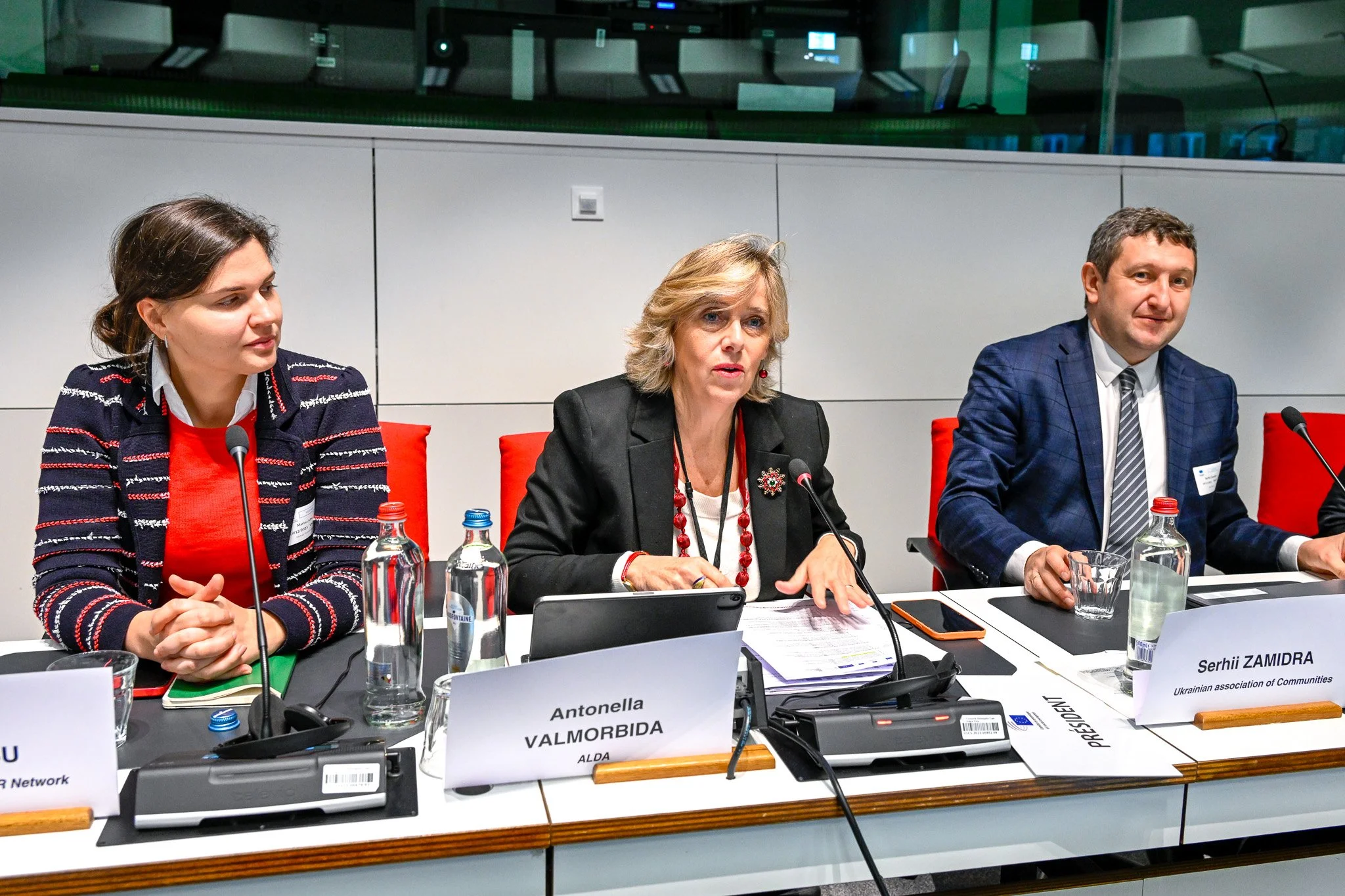 Trois personnes assises à une table de conférence avec des micros et des bouteilles d'eau, participant à une réunion officielle.