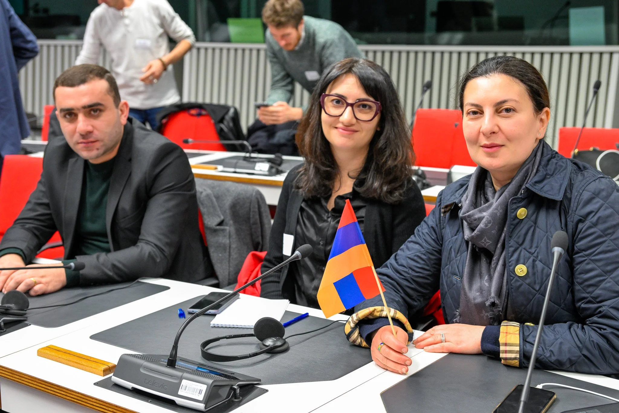 Un groupe de personnes assises à une table de conférence, tenant un drapeau arménien, avec des microphones devant eux.