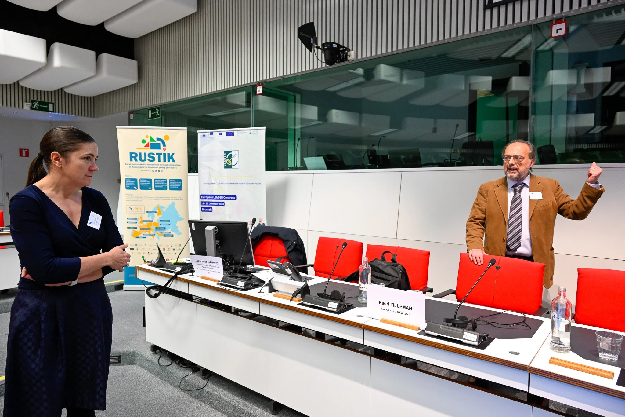 Deux personnes discutent dans une salle de conférence moderne, avec des affiches de projets intitulées « RUSTIK » et « European LEADER Congress ». Des chaises rouges et des ordinateurs sont à proximité.