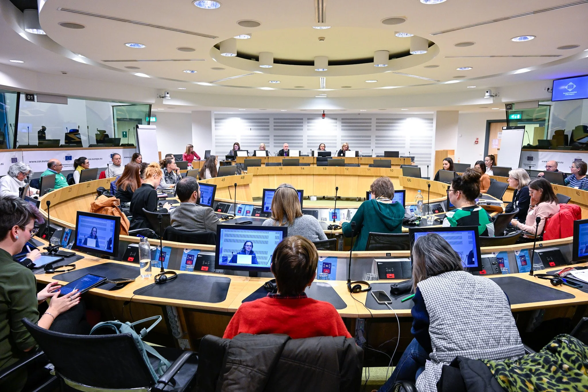 Réunion en salle de conférence avec plusieurs personnes autour d'une table en forme d'arc, utilisant des ordinateurs et des écrans devant elles, dans un environnement professionnel. Présence de micros et d'équipements audiovisuels.