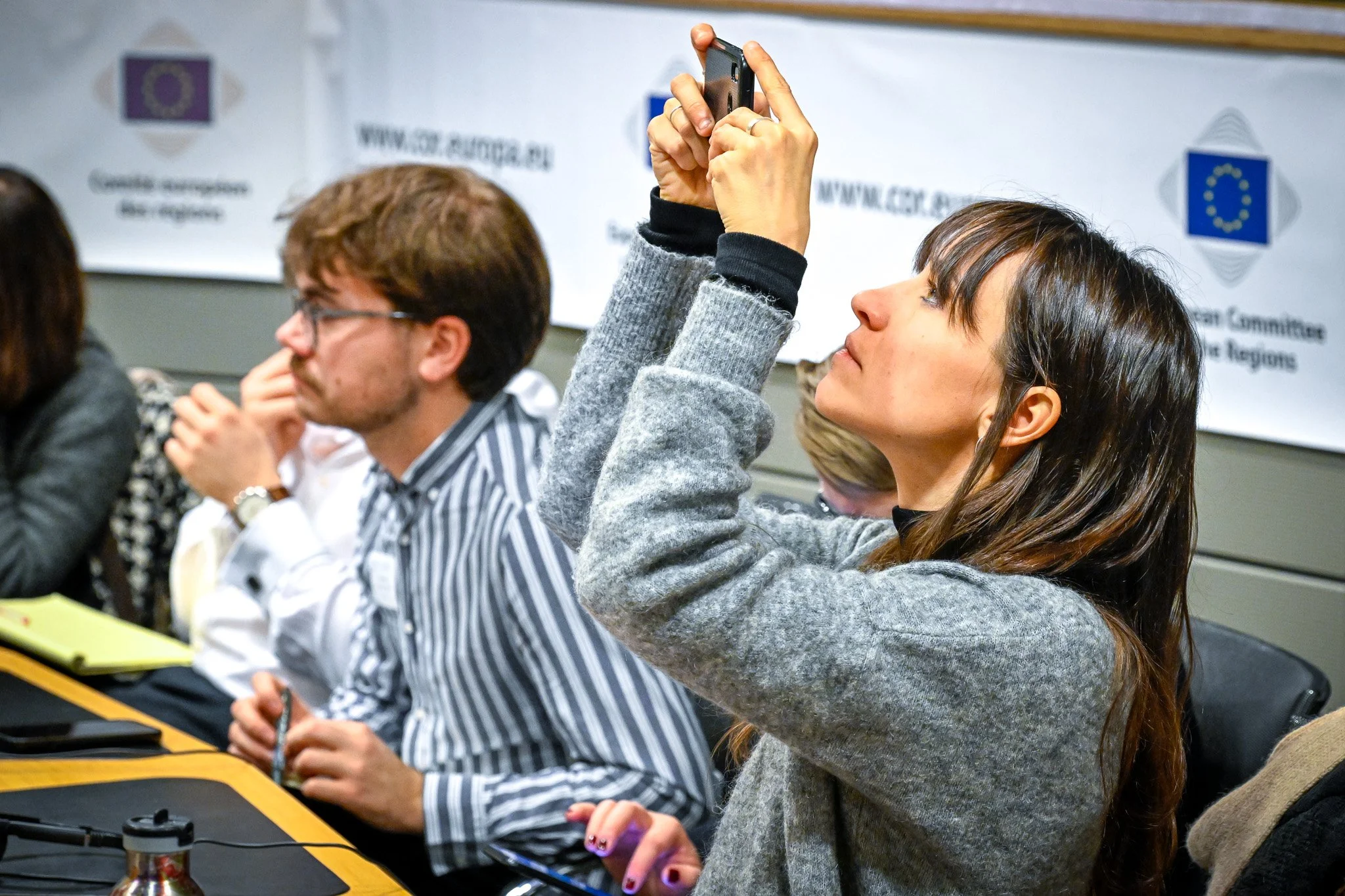 Groupe de personnes assises à une conférence, une femme prenant des photos avec un smartphone.