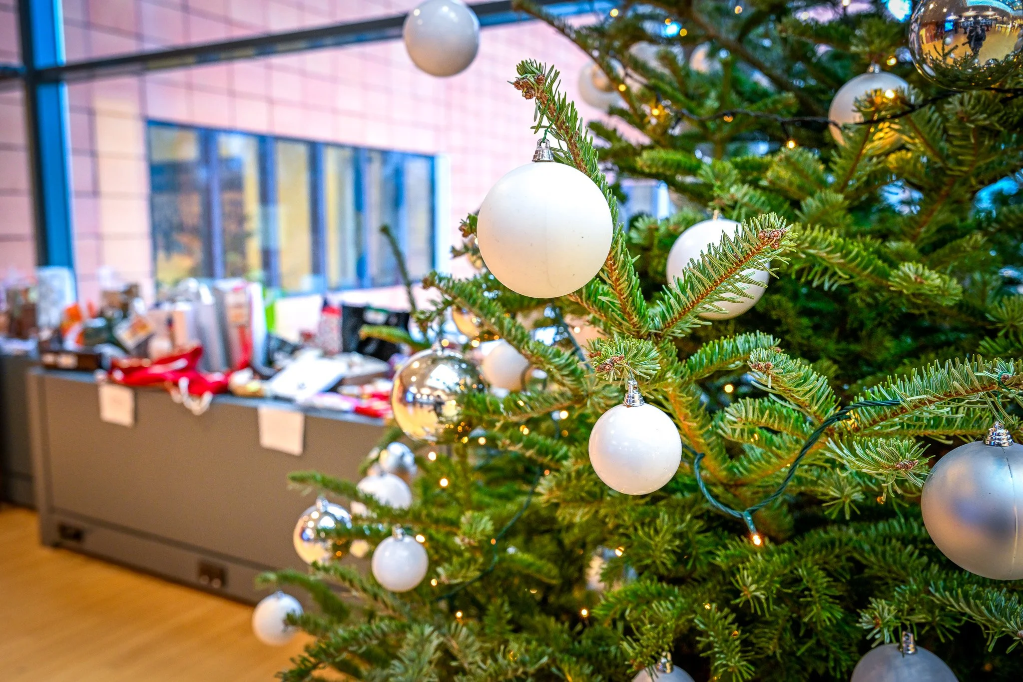 Sapin de Noël décoré avec des boules blanches et argentées, lumière de fées, à côté d'un espace de travail avec des cadeaux sur une table.
