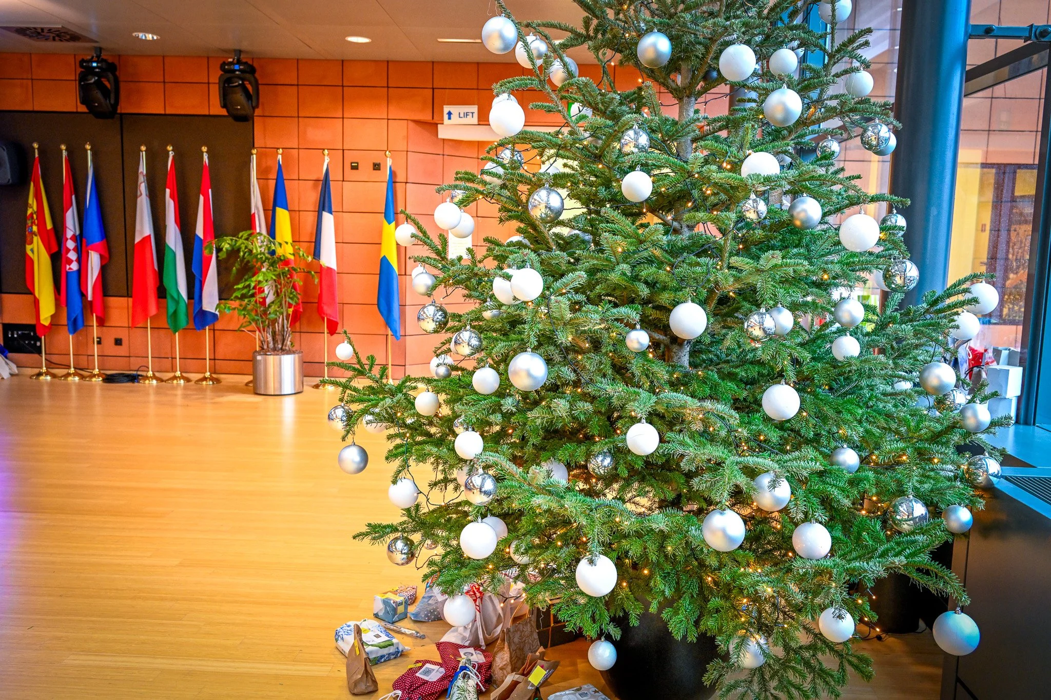 Un sapin de Noël décoré avec des boules argentées et blanches, entouré de cadeaux sur le sol, dans une salle avec plusieurs drapeaux en arrière-plan.