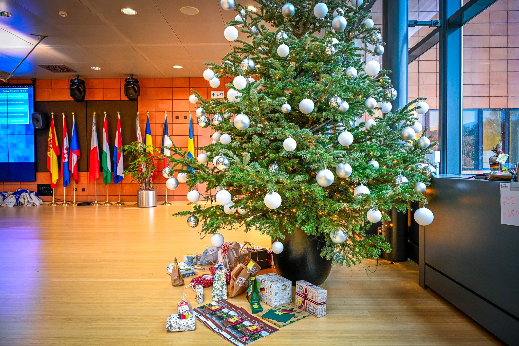 Sapin de Noël décoré avec des boules blanches et argentées, entouré de cadeaux emballés, dans une salle avec des drapeaux nationaux en arrière-plan.