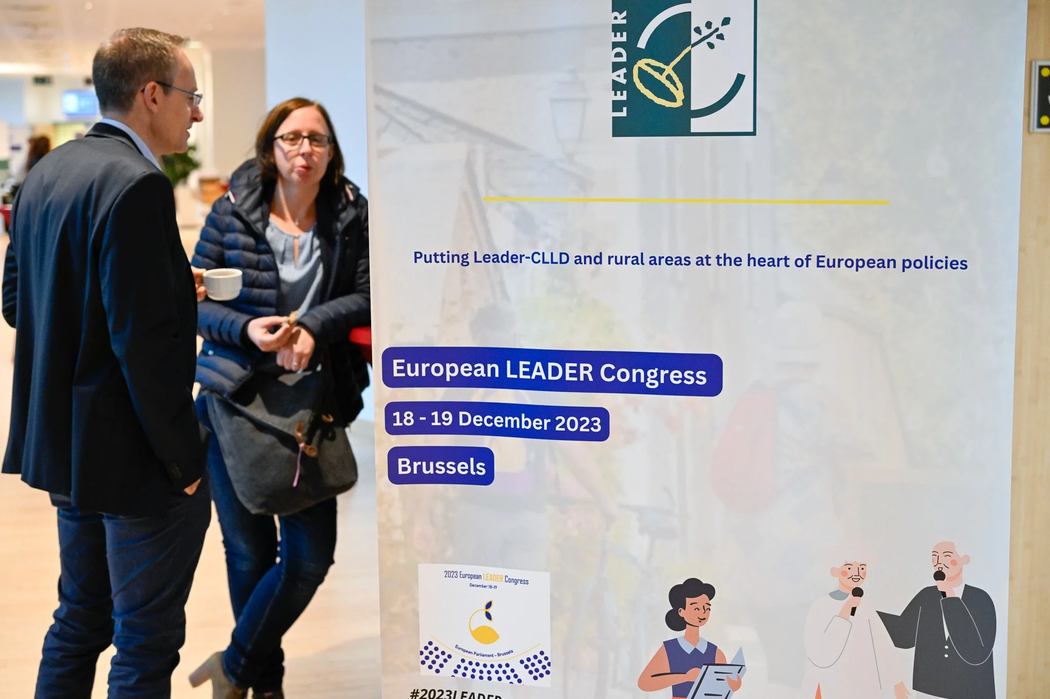 Deux personnes discutent devant une affiche pour le Congrès Européen LEADER qui a lieu les 18 et 19 décembre 2023 à Bruxelles.