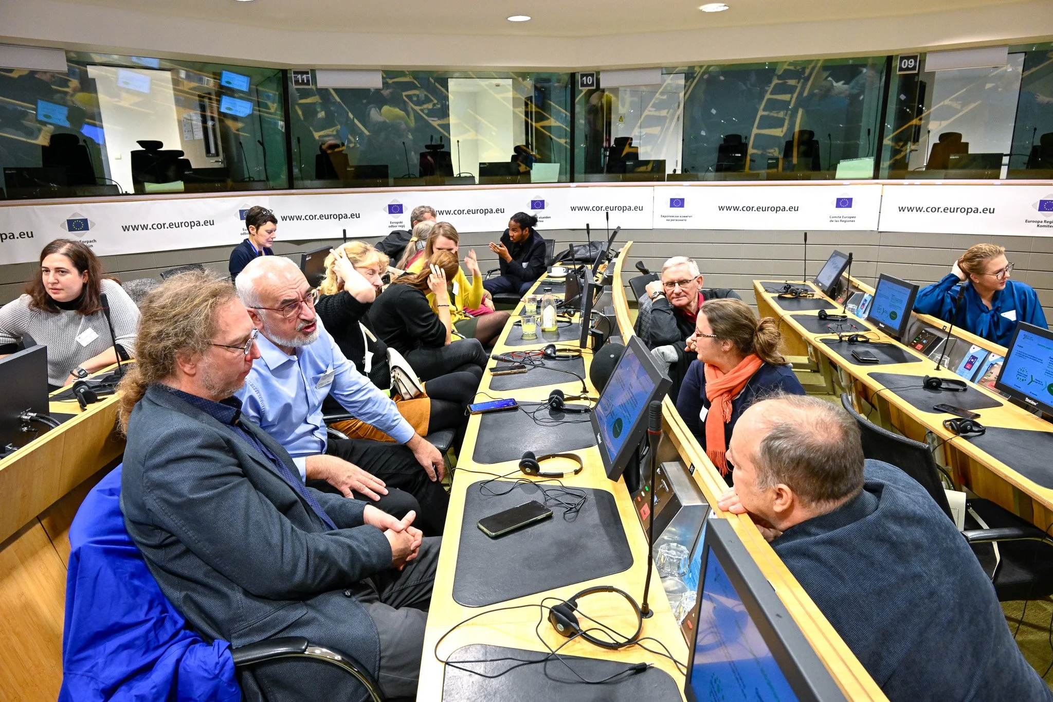Des personnes en discussion dans une salle de conférence avec des ordinateurs et écrans, logo cor.europa.eu sur les murs.