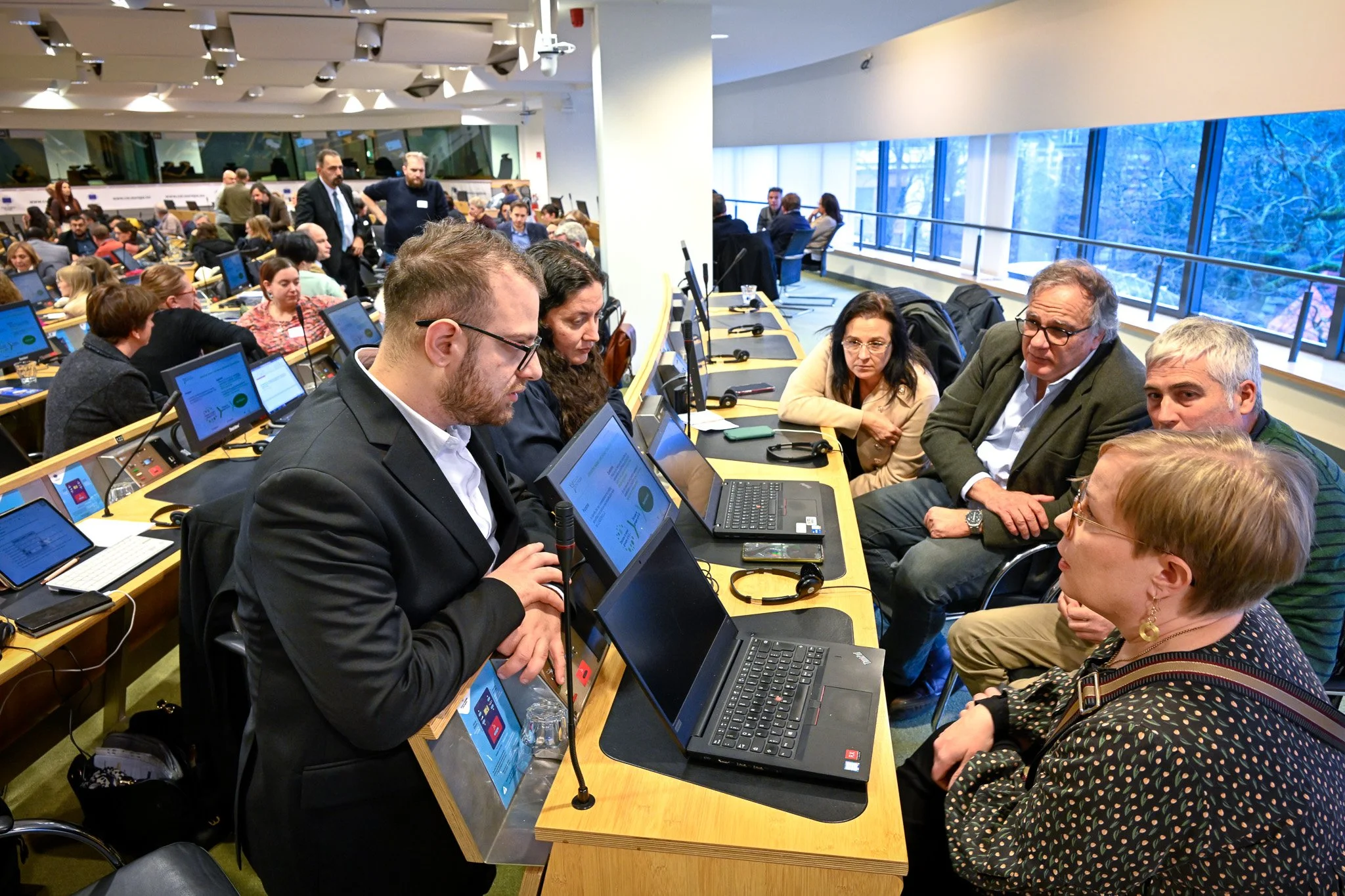 Une salle de conférence où des personnes discutent autour de ordinateurs portables, avec des rangées de participants en arrière-plan.