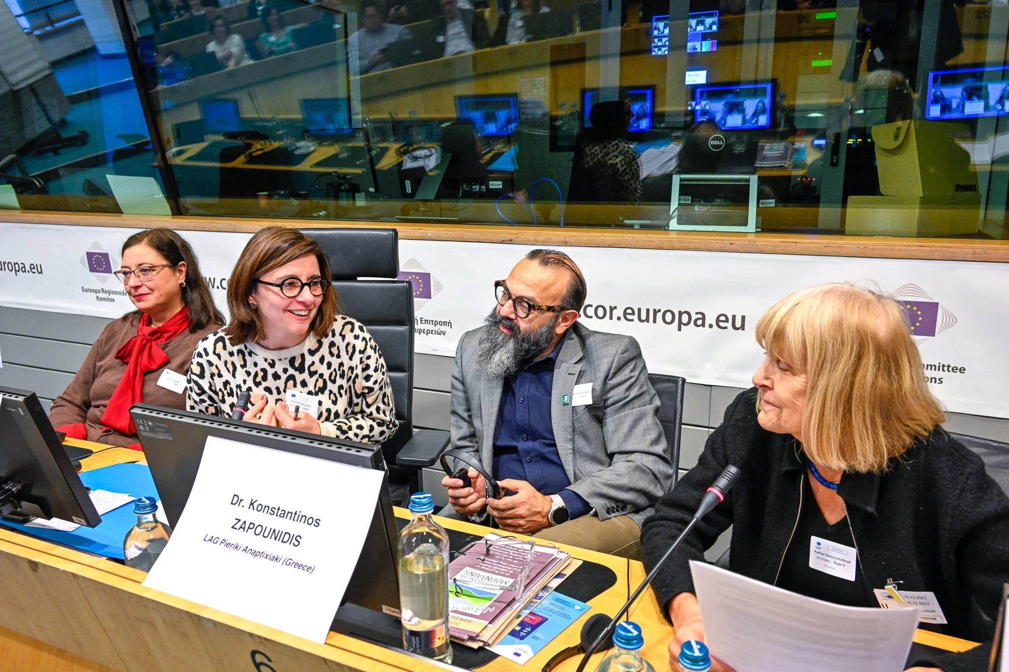 Quatre personnes assises à une table lors d'une conférence au sein du Comité européen des régions, discutant et utilisant des ordinateurs ; fond avec écran et vitres séparant les délégués; panneau indiquant Dr. Konstantinos ZARPOUNIDIS. Bouteilles d'