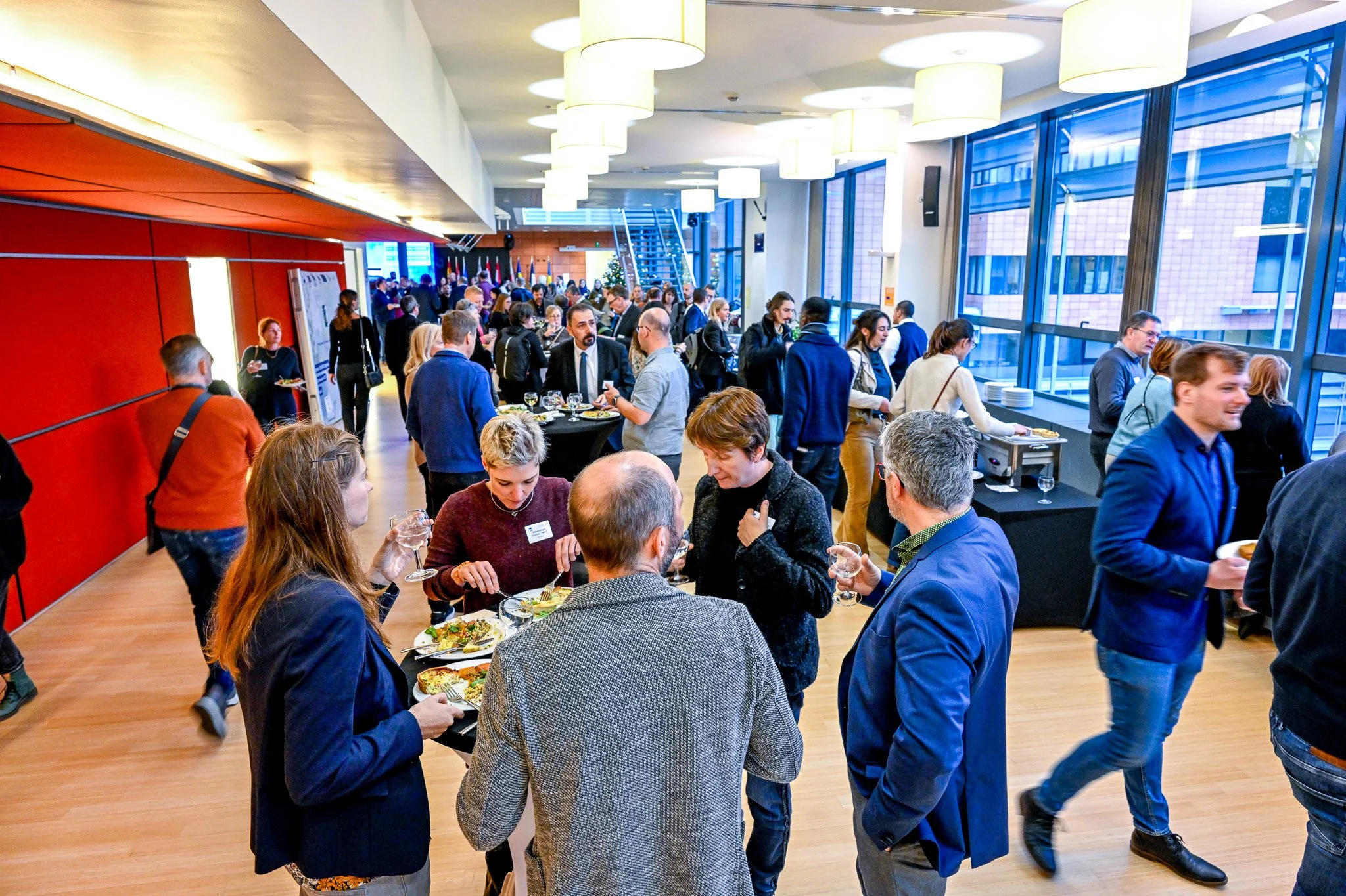 Un groupe de personnes participe à un événement social dans une salle moderne avec des fenêtres. Plusieurs personnes sont debout, discutant et mangeant autour de tables hautes avec de la nourriture et des boissons."