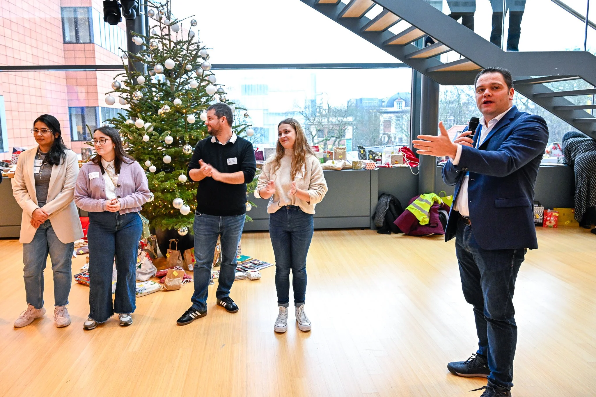 Un groupe de personnes debout à côté d'un sapin de Noël décoré, dans une pièce lumineuse avec un sol en bois, une grande fenêtre et des escaliers. Une personne parle avec un micro.