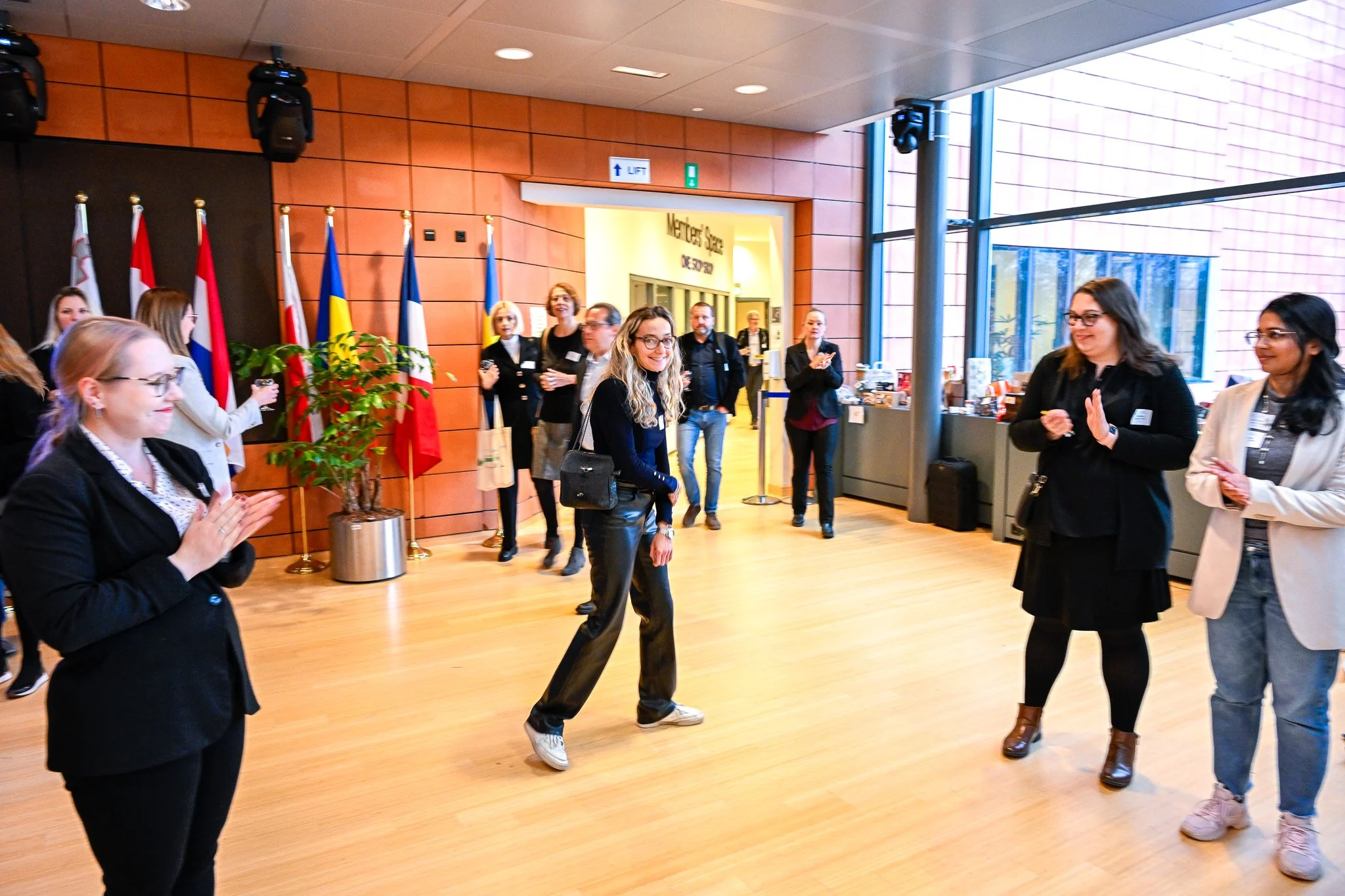 Un groupe de personnes dans un hall avec plusieurs drapeaux en arrière-plan, applaudissant une femme au centre. Le sol est en bois clair, et il y a des fenêtres sur le côté droit.