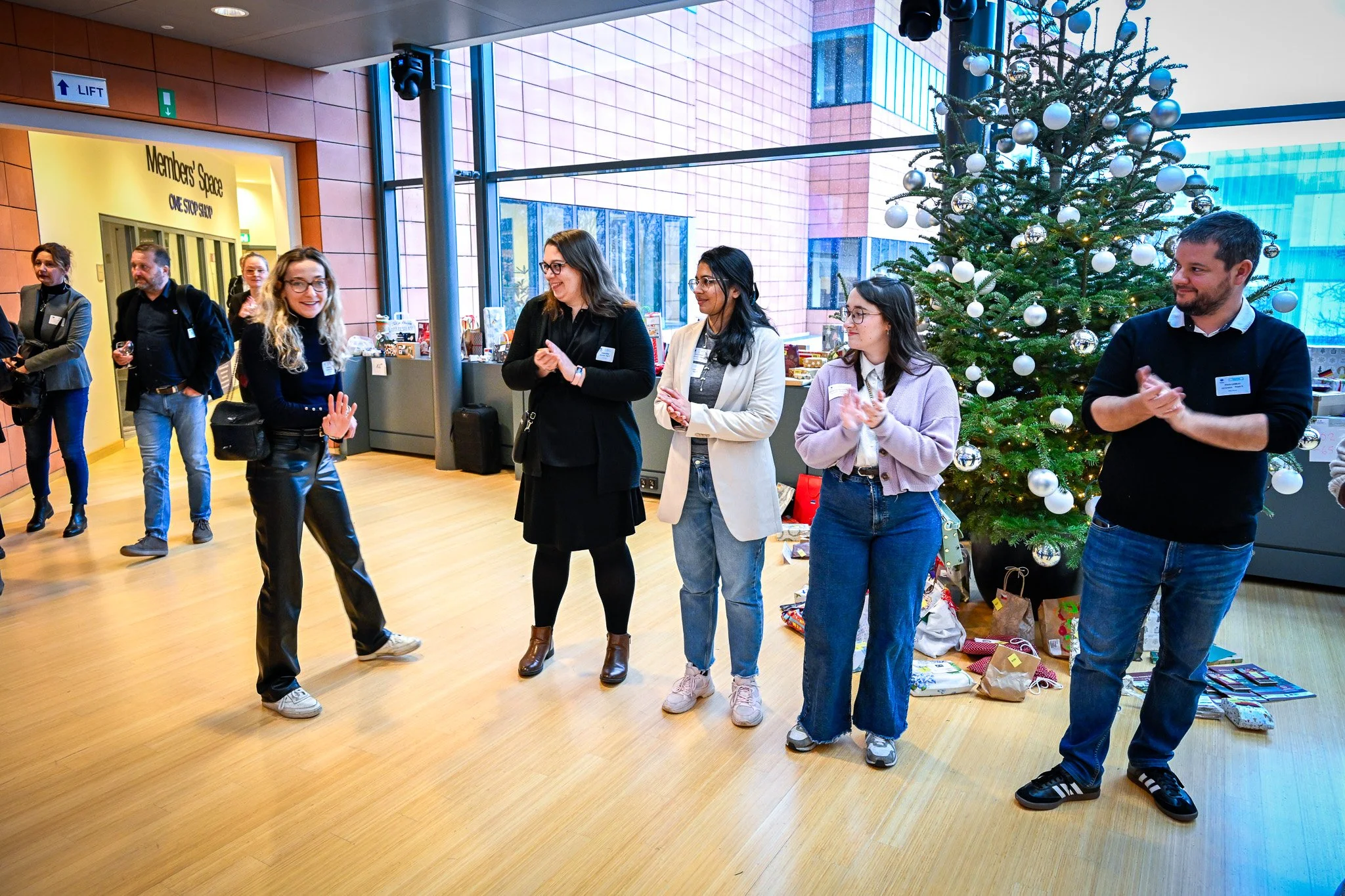 Groupe de personnes debout dans une pièce décorée pour Noël avec un sapin, discutant et applaudi.
