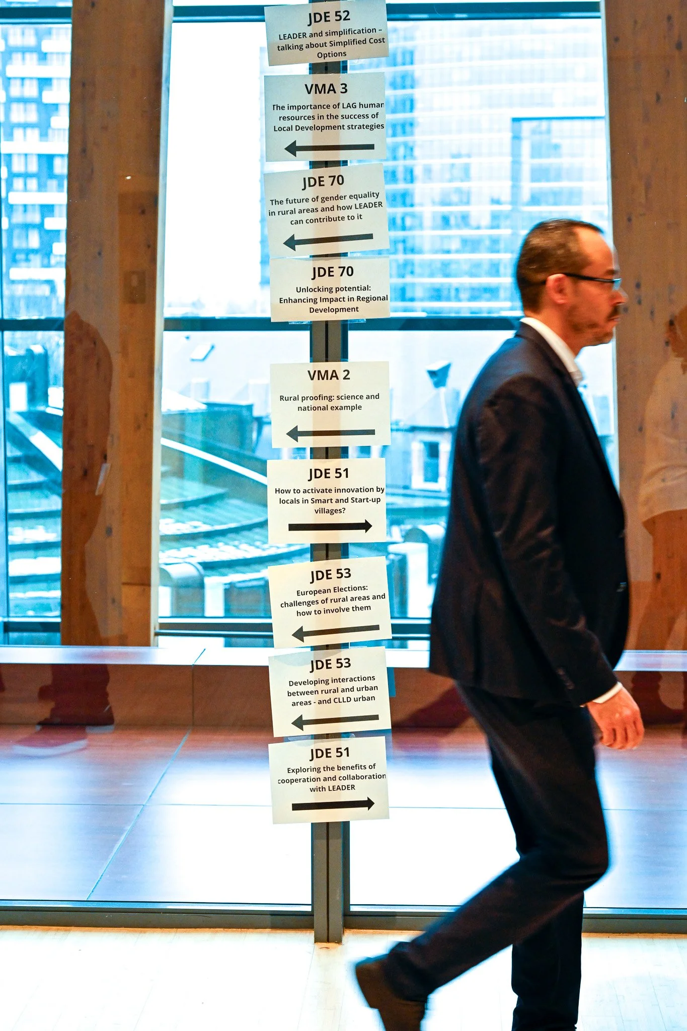 Un homme en costume marche devant une série d'affiches sur des thèmes de développement régional, dans un bâtiment moderne avec de grandes fenêtres.