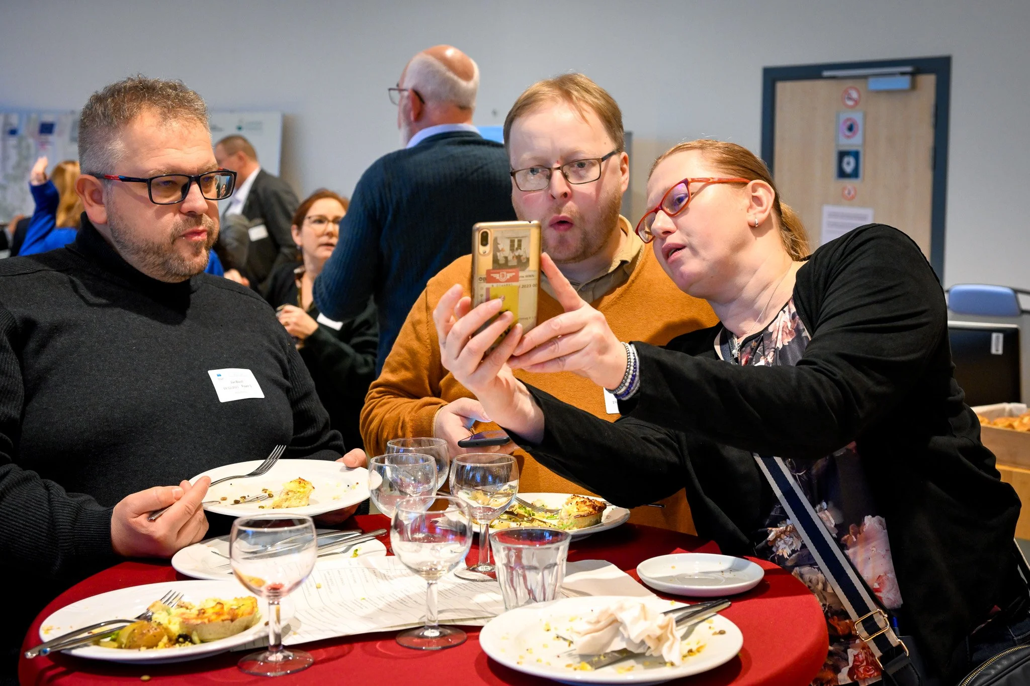 Des personnes assises à une table lors d'un événement, partageant un moment autour d'un téléphone portable, avec des assiettes de nourriture devant elles.