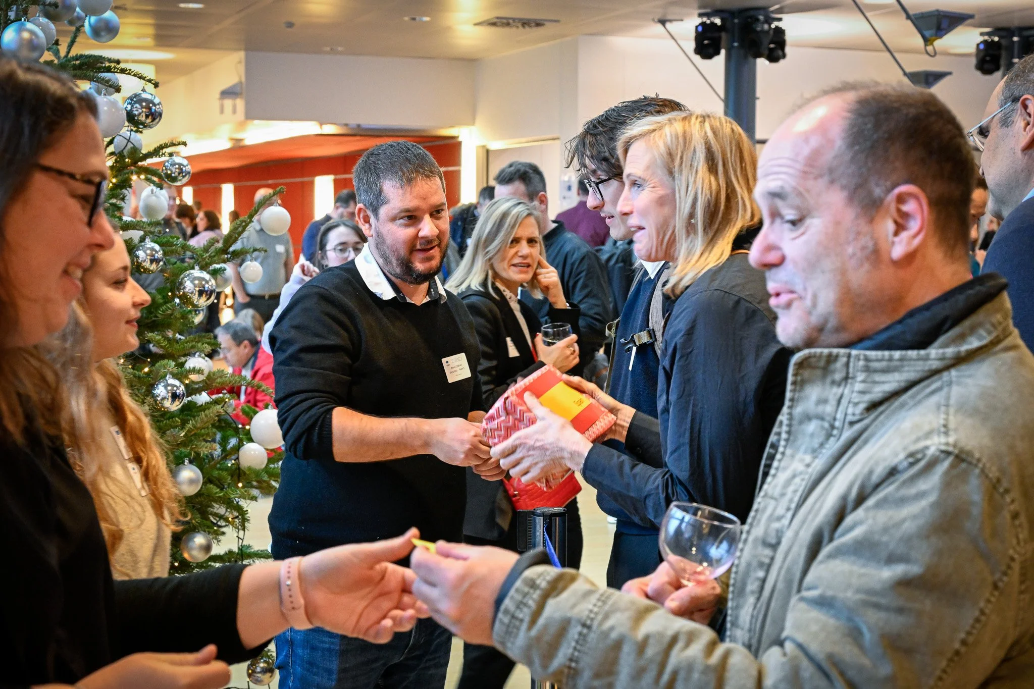 Groupe de personnes échangeant des cadeaux lors d'un événement de Noël avec un sapin décoré en arrière-plan.