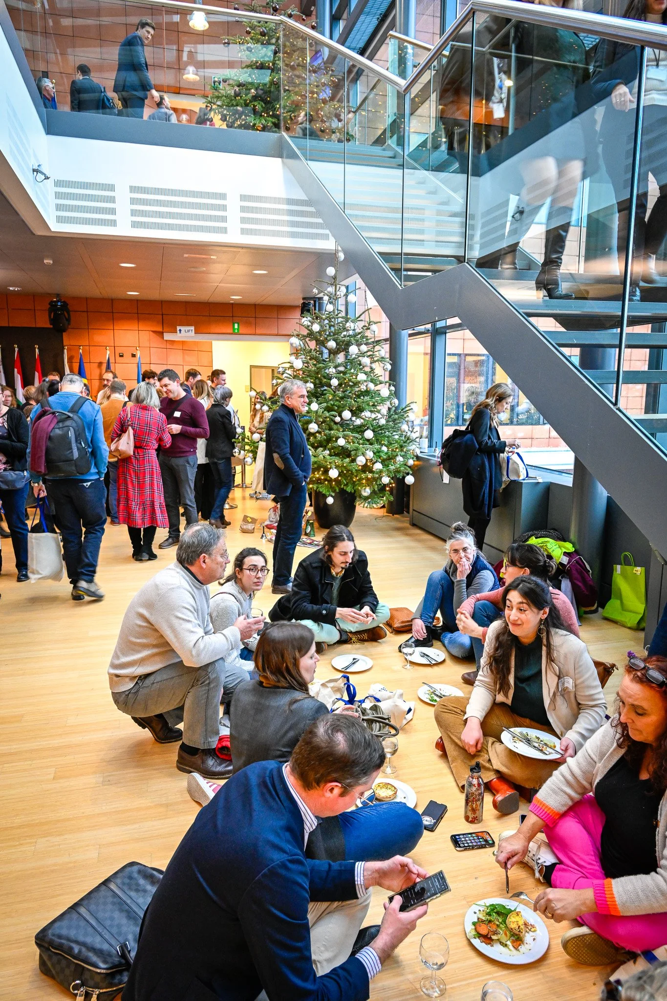 Groupe de personnes assises par terre dans un hall avec un sapin de Noël décoré, mangeant et discutant. D'autres personnes se tiennent debout à l'arrière-plan près des drapeaux. Un escalier en verre est visible.