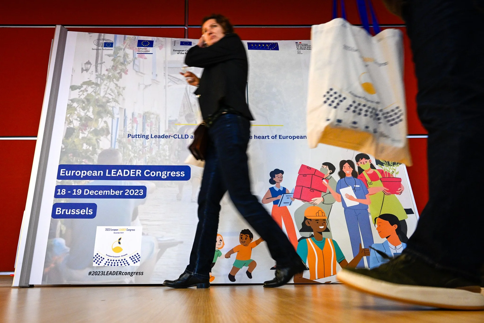 Banque d'information du Congrès LEADER européen 2023 à Bruxelles. Un passant flou devant une affiche avec illustrations de travailleurs communautaires dans un style plat. Texte indiquant les dates du congrès : 18-19 décembre 2023.