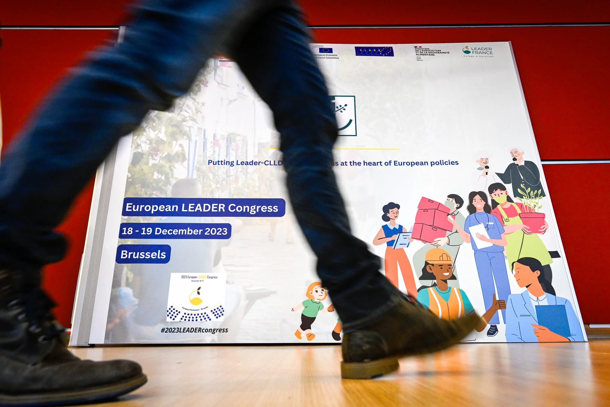Personne marchant devant une affiche du Congrès européen LEADER à Bruxelles, 18-19 décembre 2023, avec illustrations de divers professionnels.