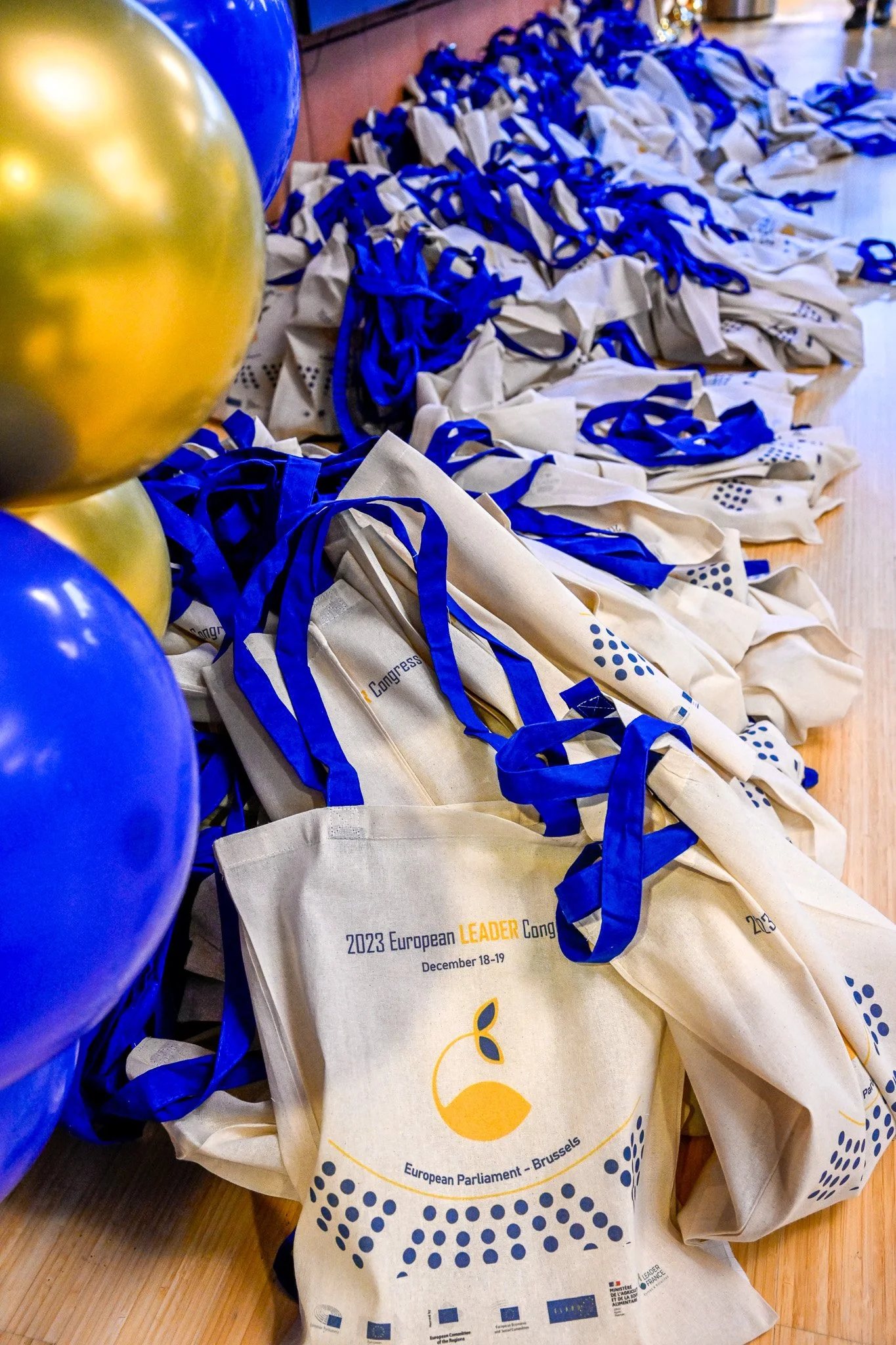 Photographie d'événement à Bruxelles Sacs en toile beige avec anses bleues et texte "2023 European LEADER Congress" au Parlement Européen, Bruxelles. Des ballons dorés et bleus sont visibles à gauche.