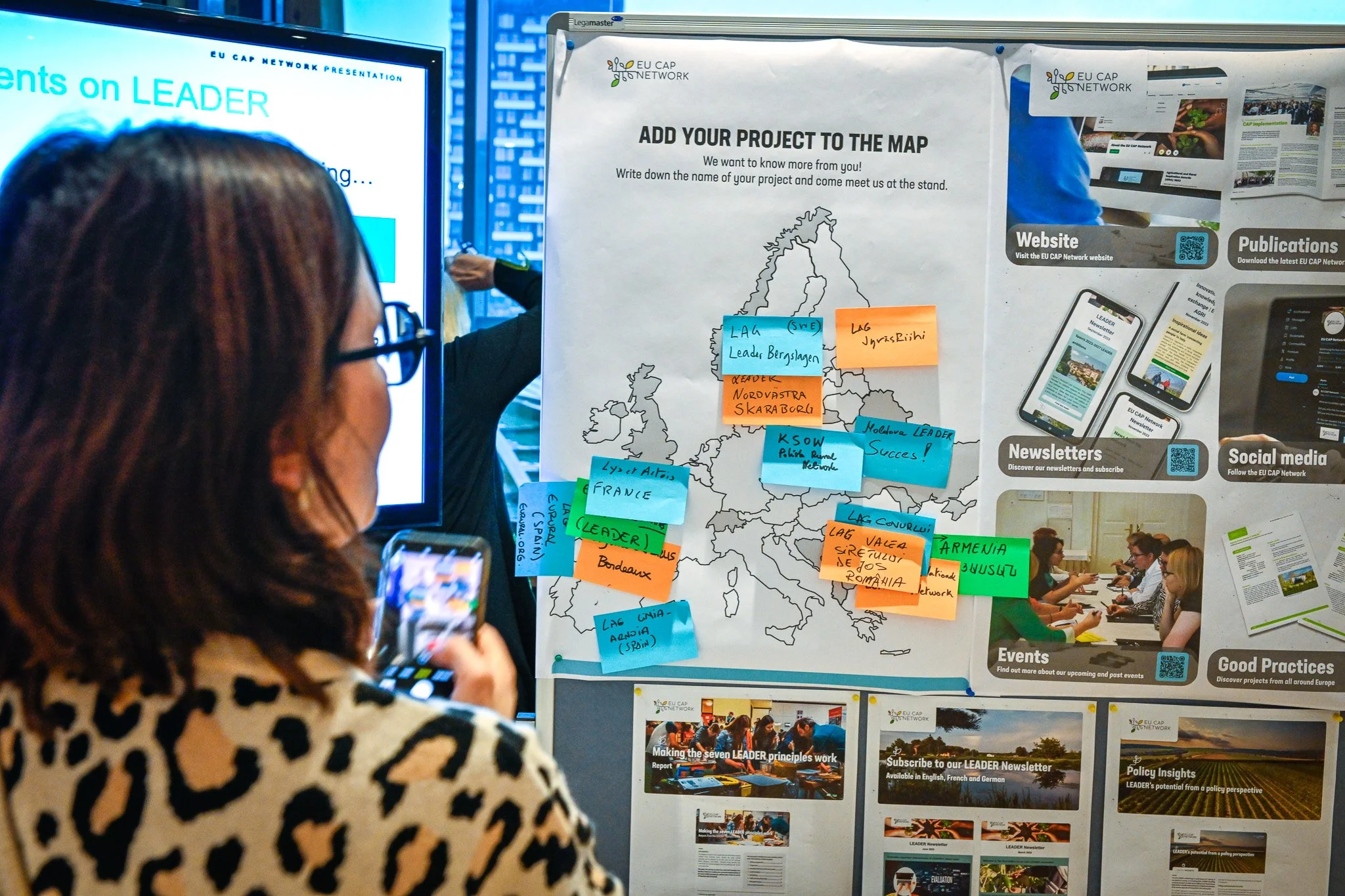 Personne regardant une carte de l'Europe avec des notes autocollantes représentant des projets du réseau EU CAP, entourée de matériel informatif sur les publications, les réseaux sociaux et les événements.