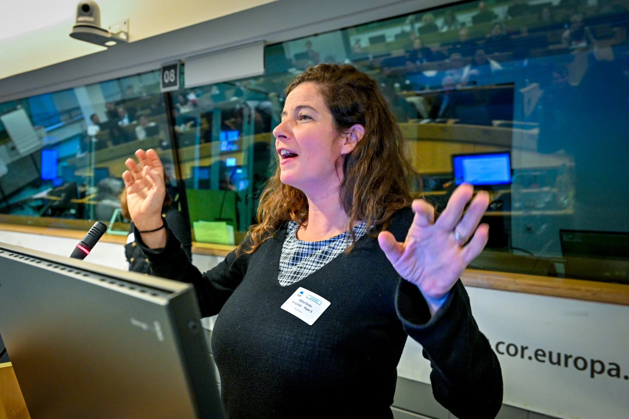 Une femme parle devant un pupitre dans un environnement de conférence, avec un écran d'ordinateur et un micro visibles. Elle porte une étiquette nominative.