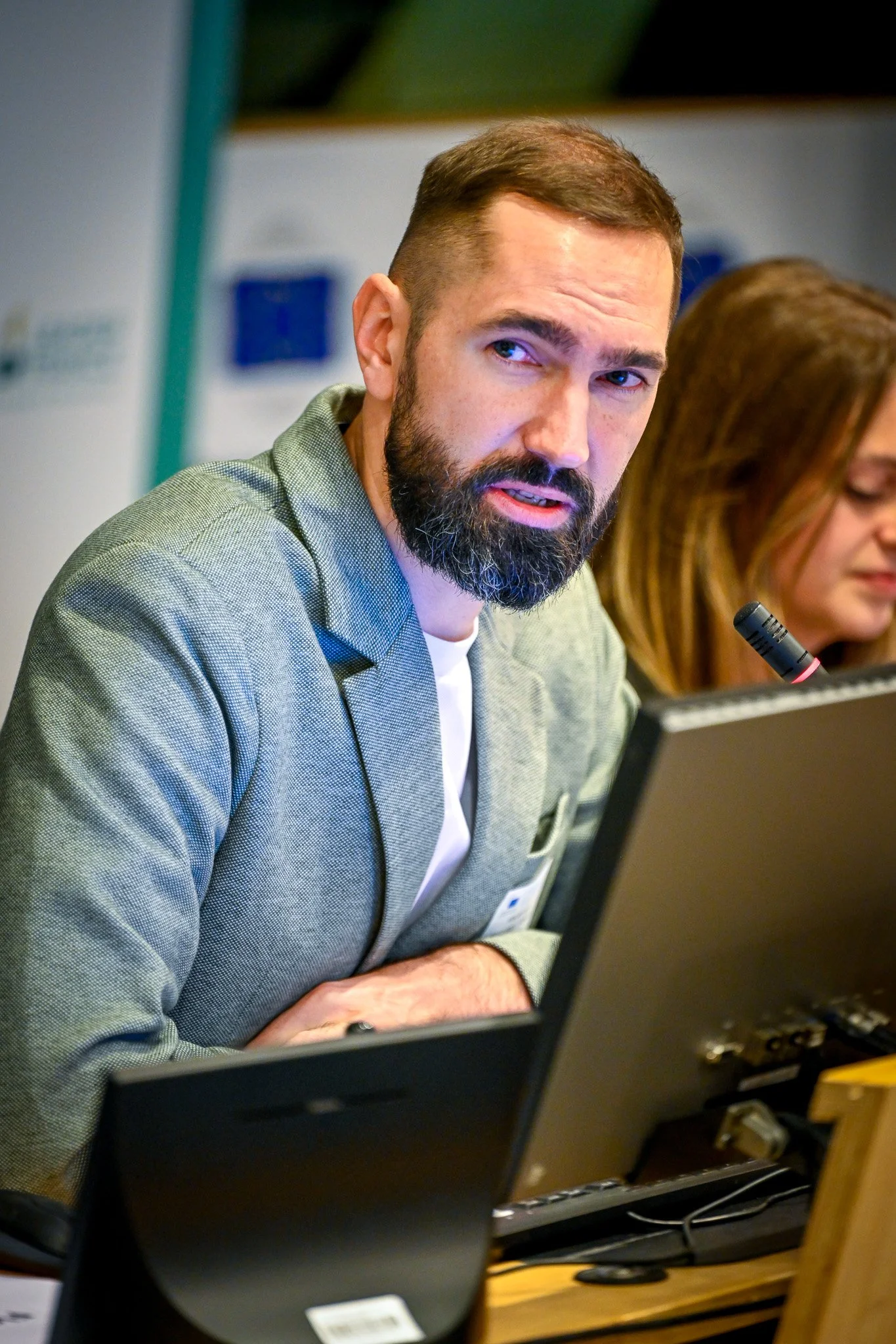 Homme barbu en costume gris parlant devant un micro, avec une femme à sa gauche, lors d'une conférence.