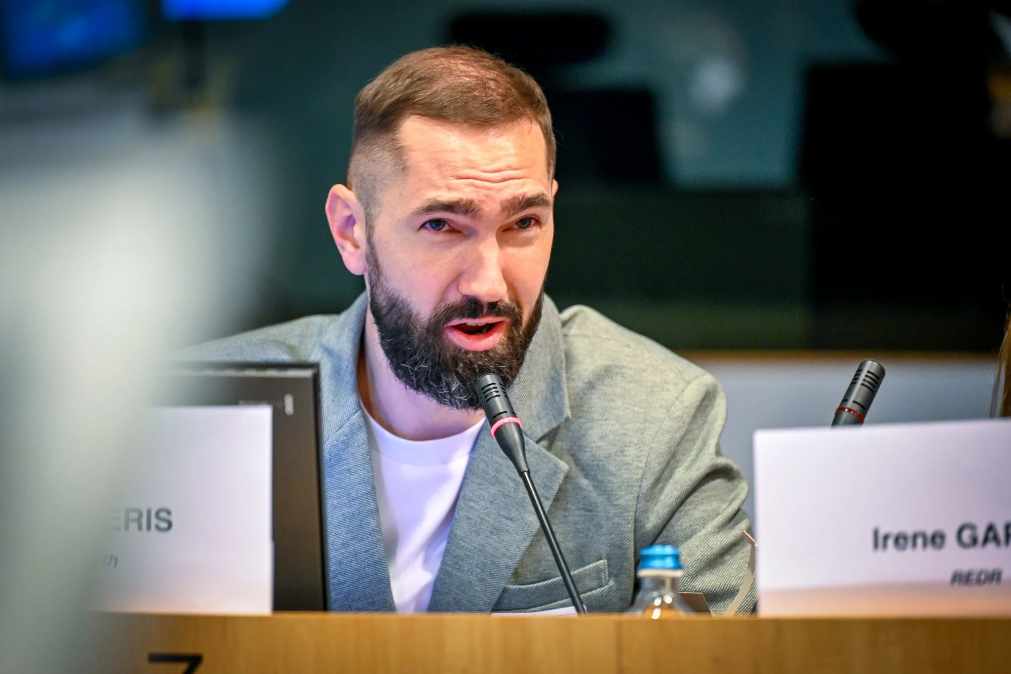 Un homme barbu en veste grise parle dans un microphone lors d'une conférence, avec un panneau de nom partiellement visible devant lui et une bouteille d'eau à côté.