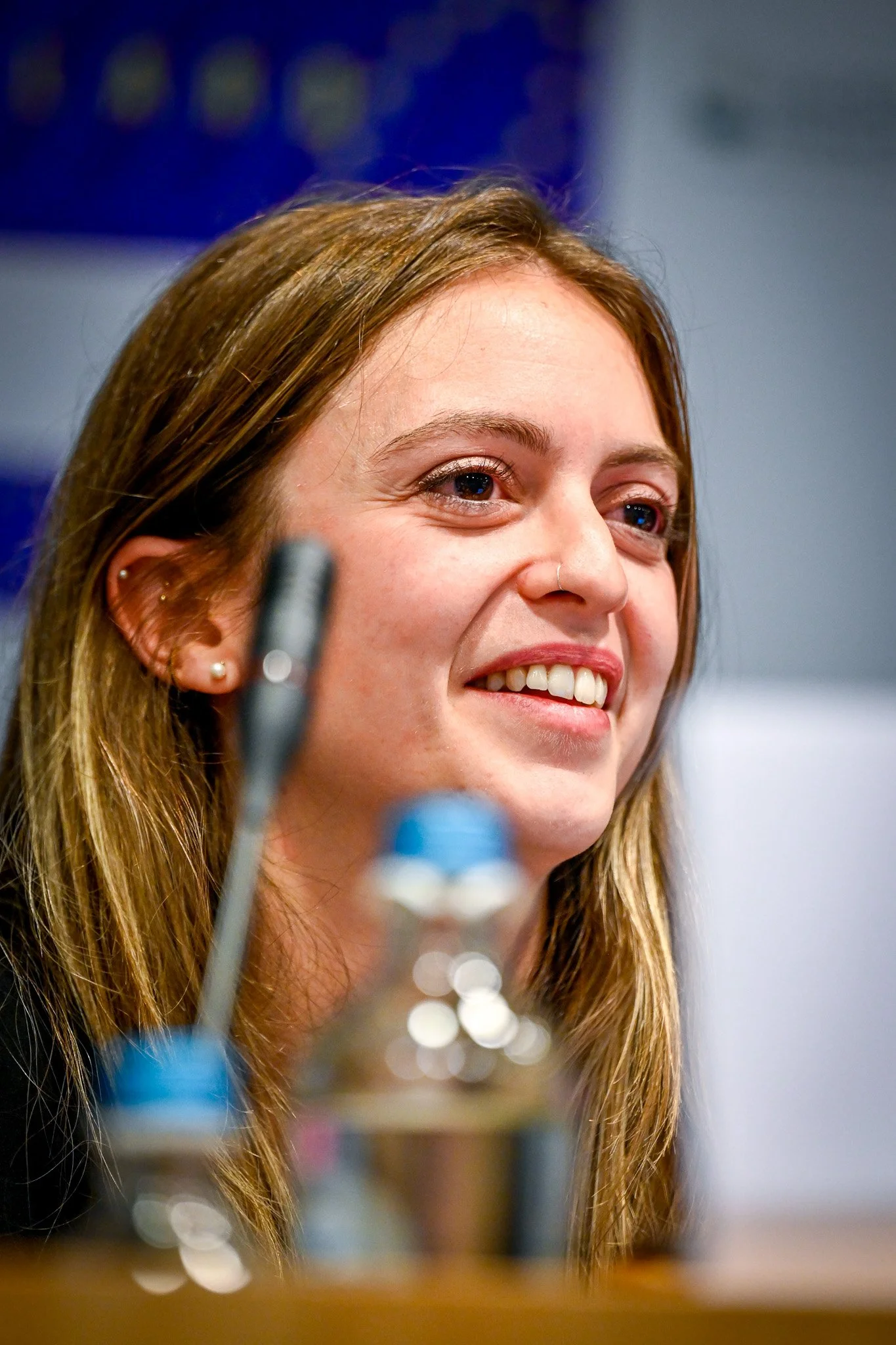 Une femme souriante lors d'une conférence, avec un micro et une bouteille d'eau floue au premier plan.