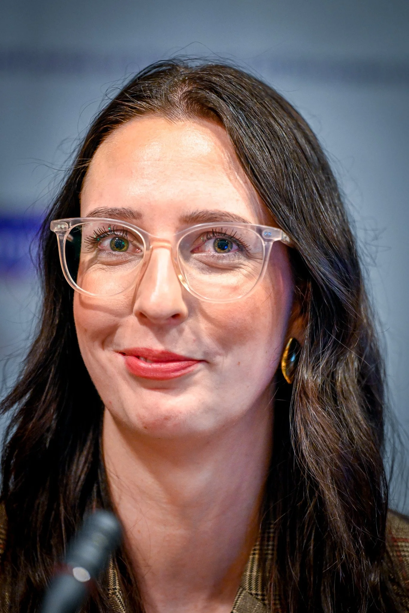 Une personne aux cheveux noirs portant des lunettes transparentes et un sourire discret.