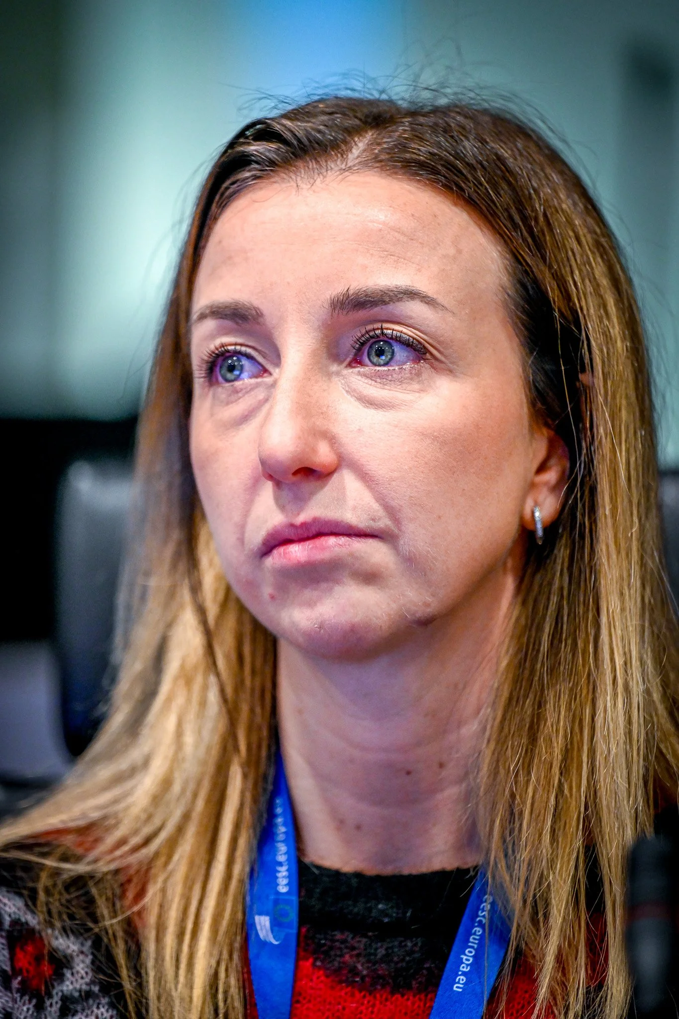 Femme avec cheveux blonds portant un badge bleu et un pull rouge, regard pensif.
