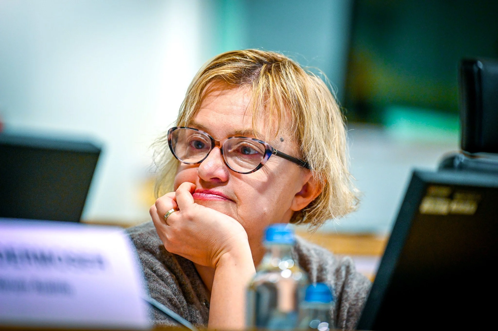 Une femme aux cheveux courts blonds portant des lunettes, appuyée sur sa main, semble pensive dans un cadre de bureau ou de conférence.