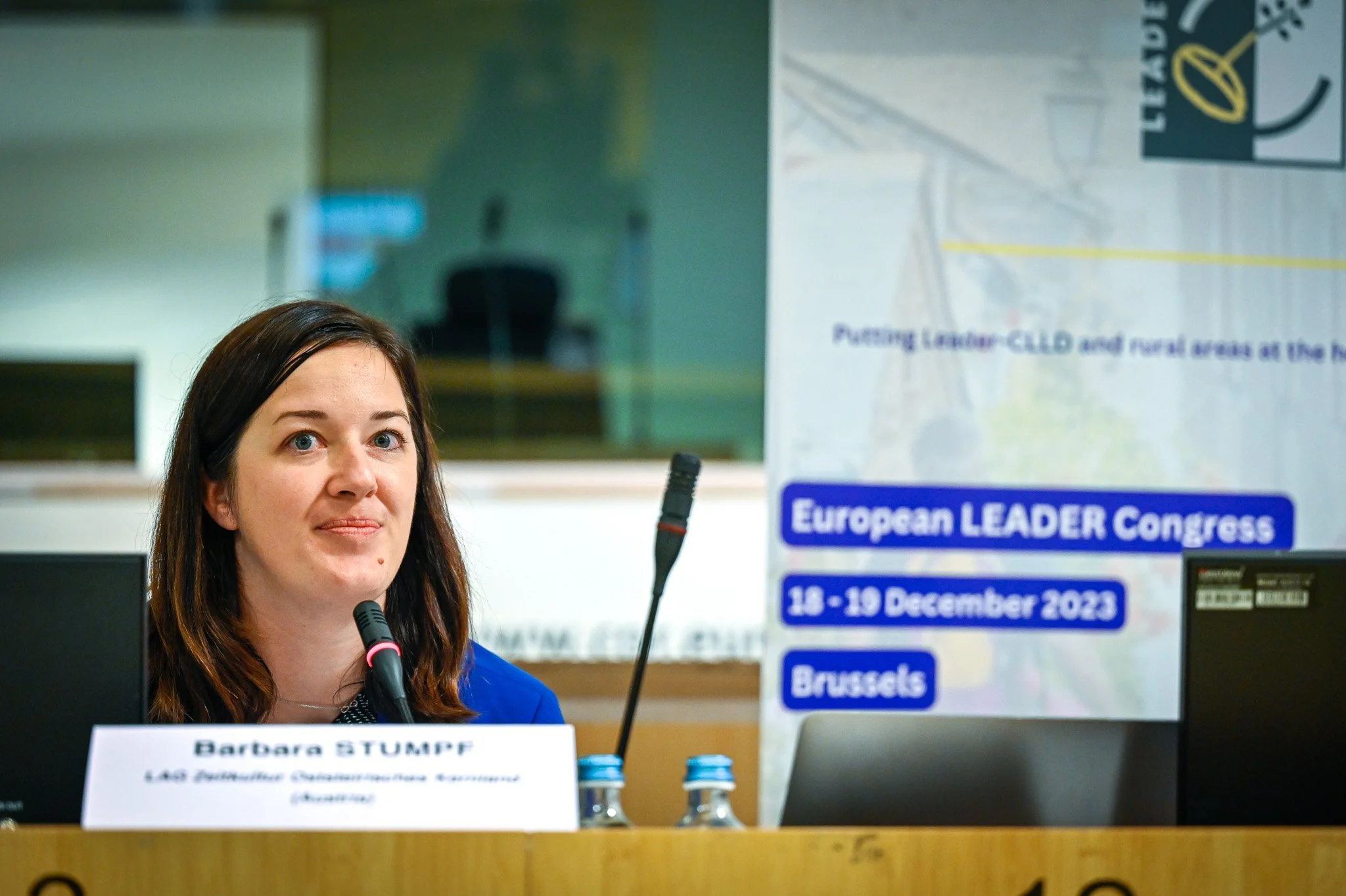 Une femme parle à une conférence, avec un panneau indiquant "European LEADER Congress, 18-19 December 2023, Brussels" en arrière-plan.
