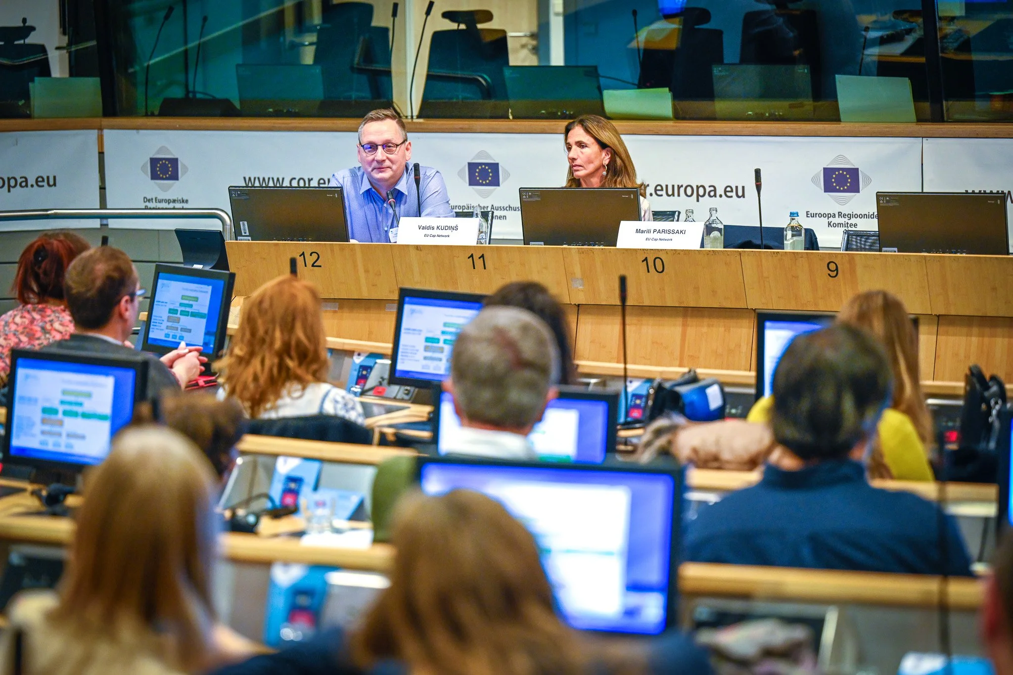 Conférence avec deux personnes sur scène devant un public utilisant des ordinateurs portables, à l'intérieur d'une salle de l'Union européenne avec des logos officiels en arrière-plan.