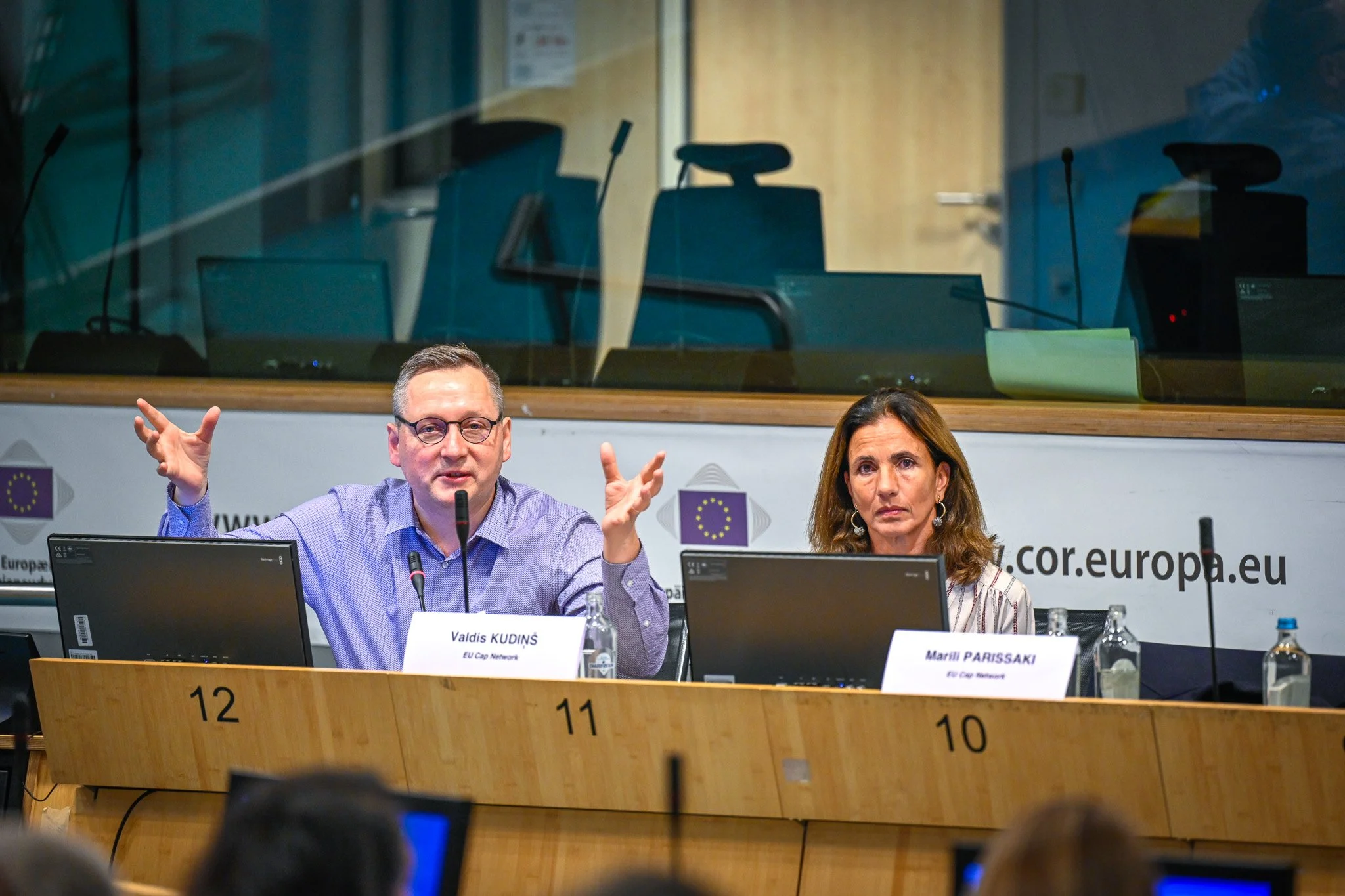 Deux personnes assises à une table de conférence avec des micros, des étiquettes de nom et des écrans d'ordinateur, lors d'une réunion ou d'un événement officiel, probablement lié à l'Union européenne.