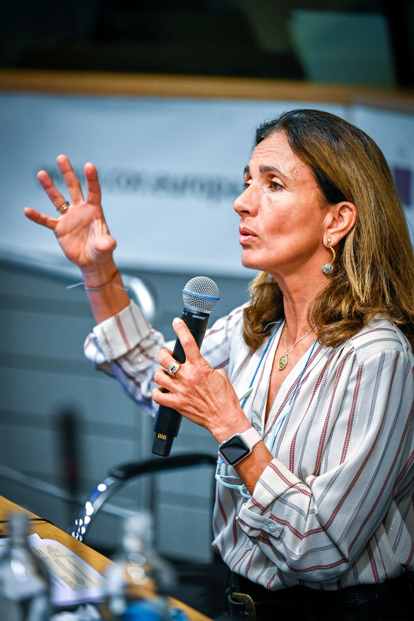 Une femme parle en tenant un microphone, gesticulant de la main gauche, portant une chemise rayée et une montre connectée.