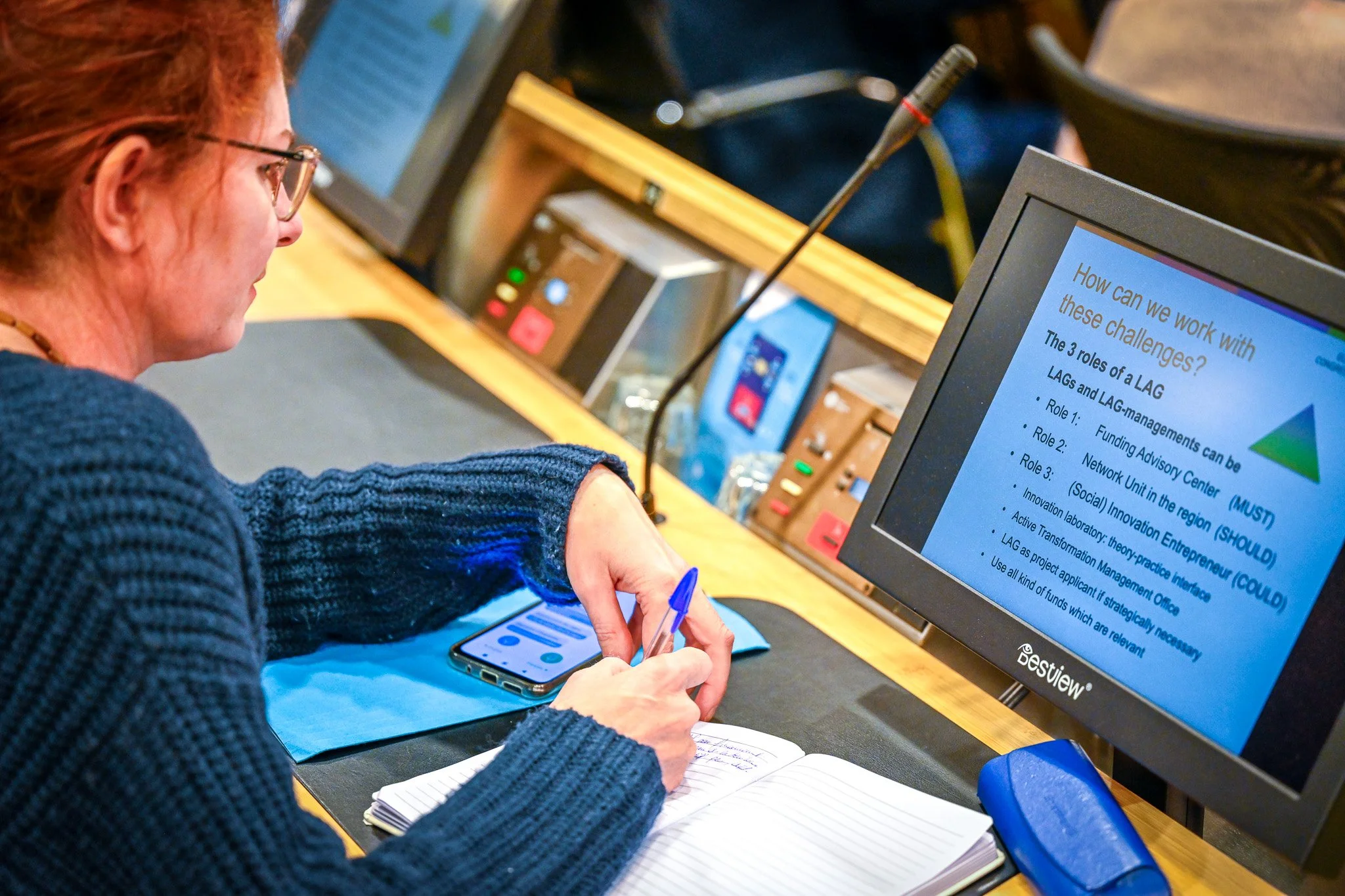 Photo d'une conférence internationale d'une agence de l'Union européenne. Une femme prend des notes lors d'une réunion, avec un écran montrant une présentation sur les rôles de la gestion LAG, et un téléphone posé sur la table.