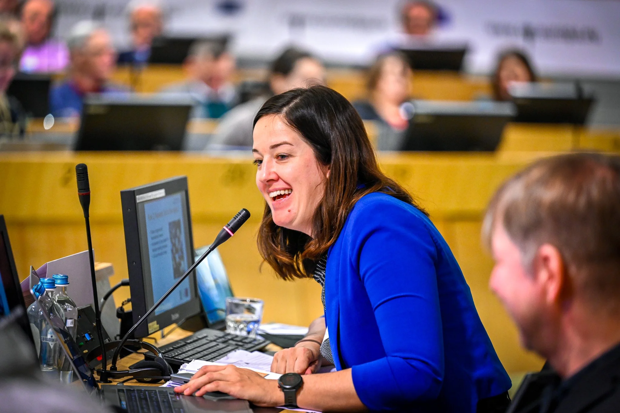 Femme souriante en train de parler dans un micro lors d'une conférence, entourée d'ordinateurs, avec un public en arrière-plan.