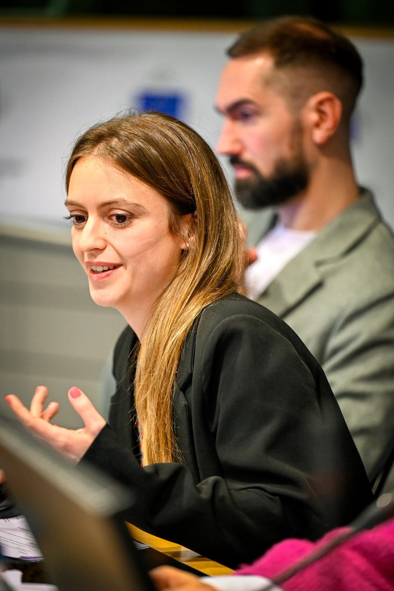Une femme parlant dans une salle de conférence, avec un homme en arrière-plan.
