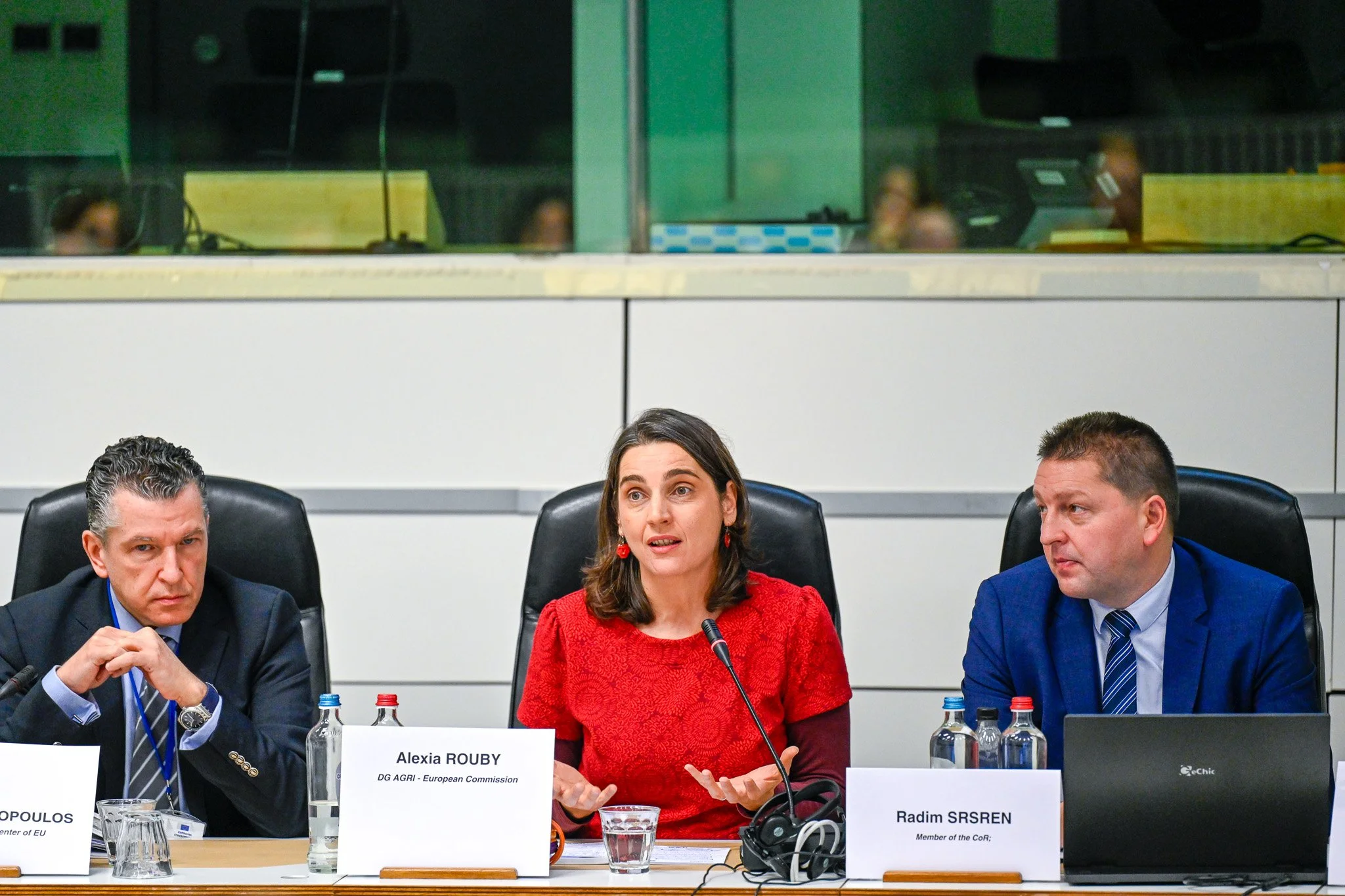 Trois personnes assises à une table de conférence avec des microphones et des étiquettes d'identification, discutant lors d'une réunion formelle.