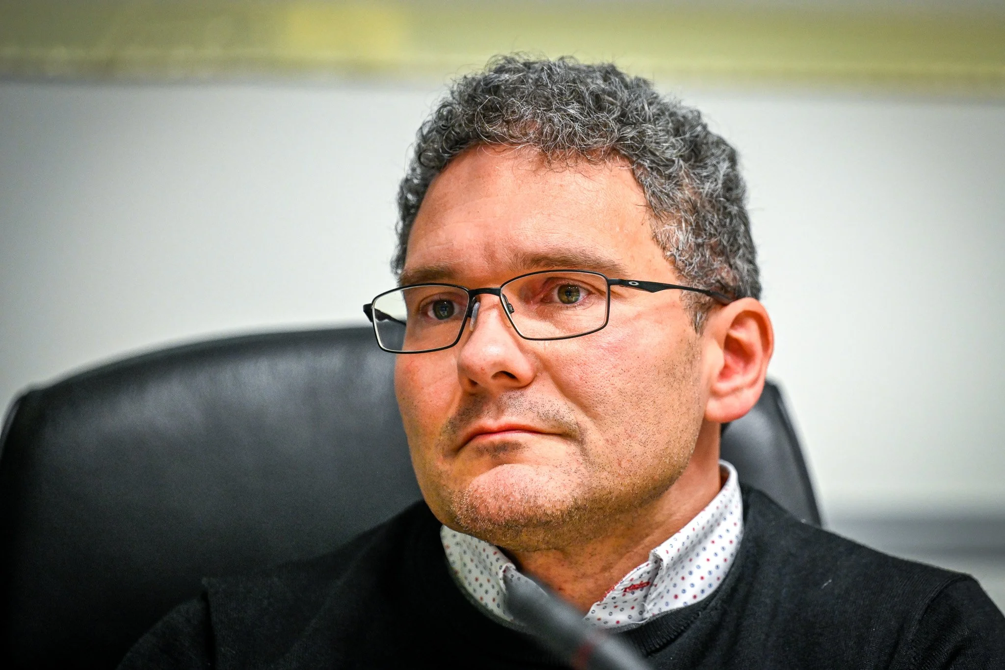 Homme aux cheveux bouclés, portant des lunettes et un pull noir, assis sur une chaise noire.