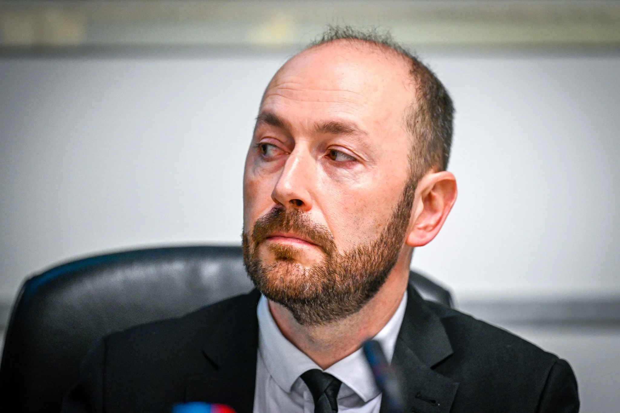 Un homme barbu, portant un costume et assis sur une chaise, regarde légèrement sur le côté.
