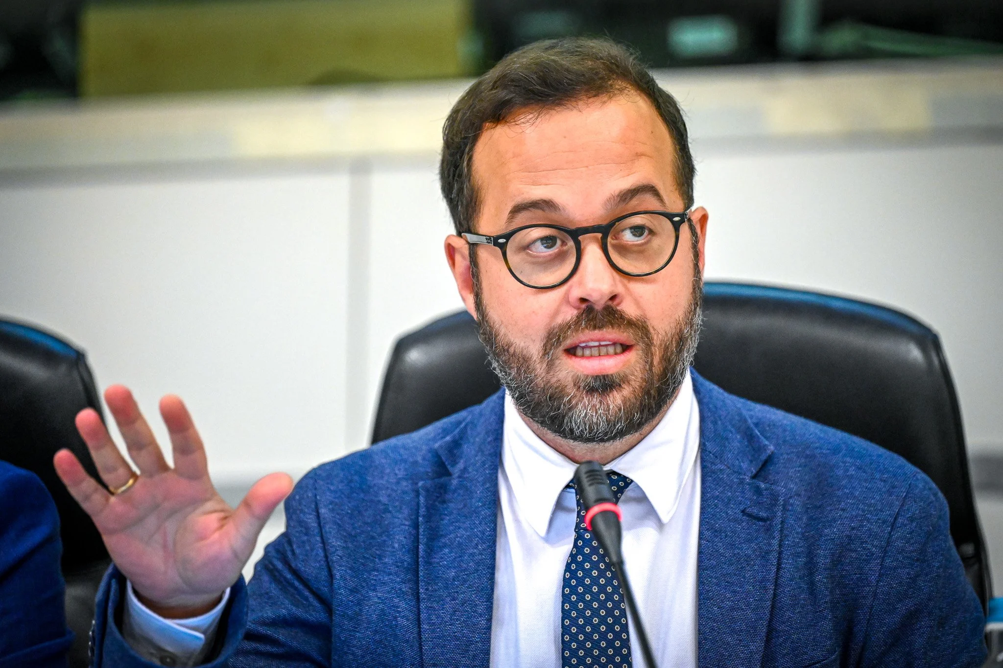 Un homme barbu avec des lunettes et un costume bleu parle dans un microphone lors d'une réunion ou d'une conférence. Sa main est levée pour accentuer son discours.