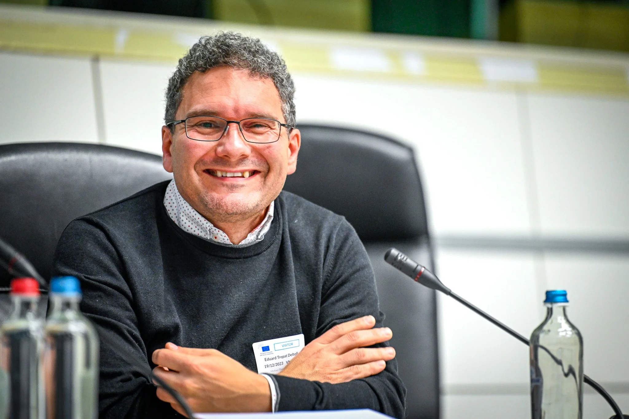 Un homme souriant portant des lunettes et un pull sombre est assis à une table avec un micro devant lui. Il y a deux bouteilles en verre sur la table. L'arrière-plan est flou, probablement dans un cadre de conférence ou de réunion.
