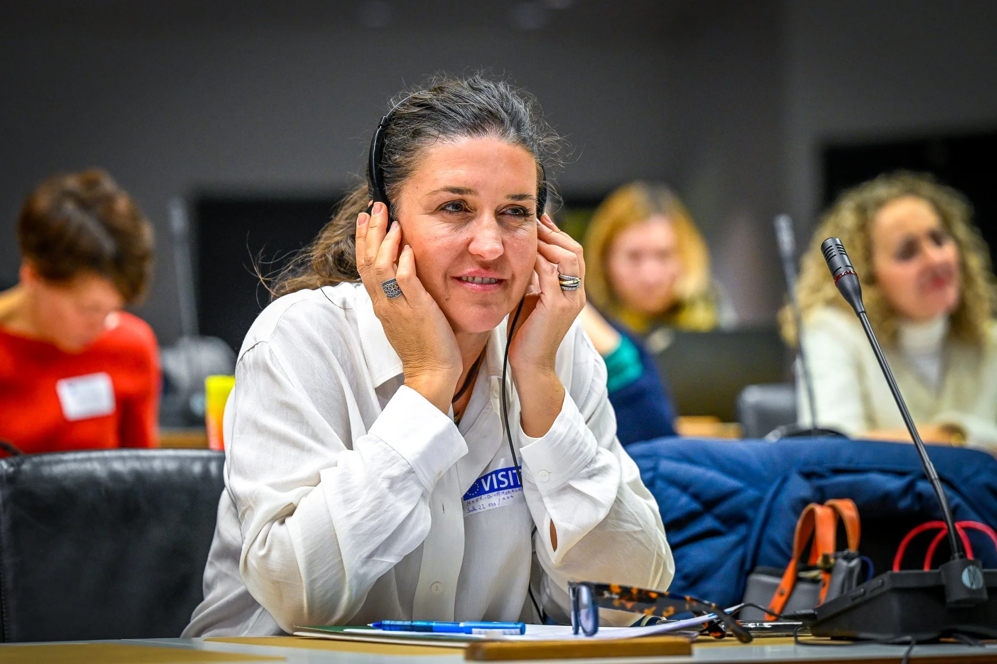 Une femme portant une chemise blanche et un casque audio, assise à une table de conférence avec un micro devant elle. D'autres personnes sont floues en arrière-plan.