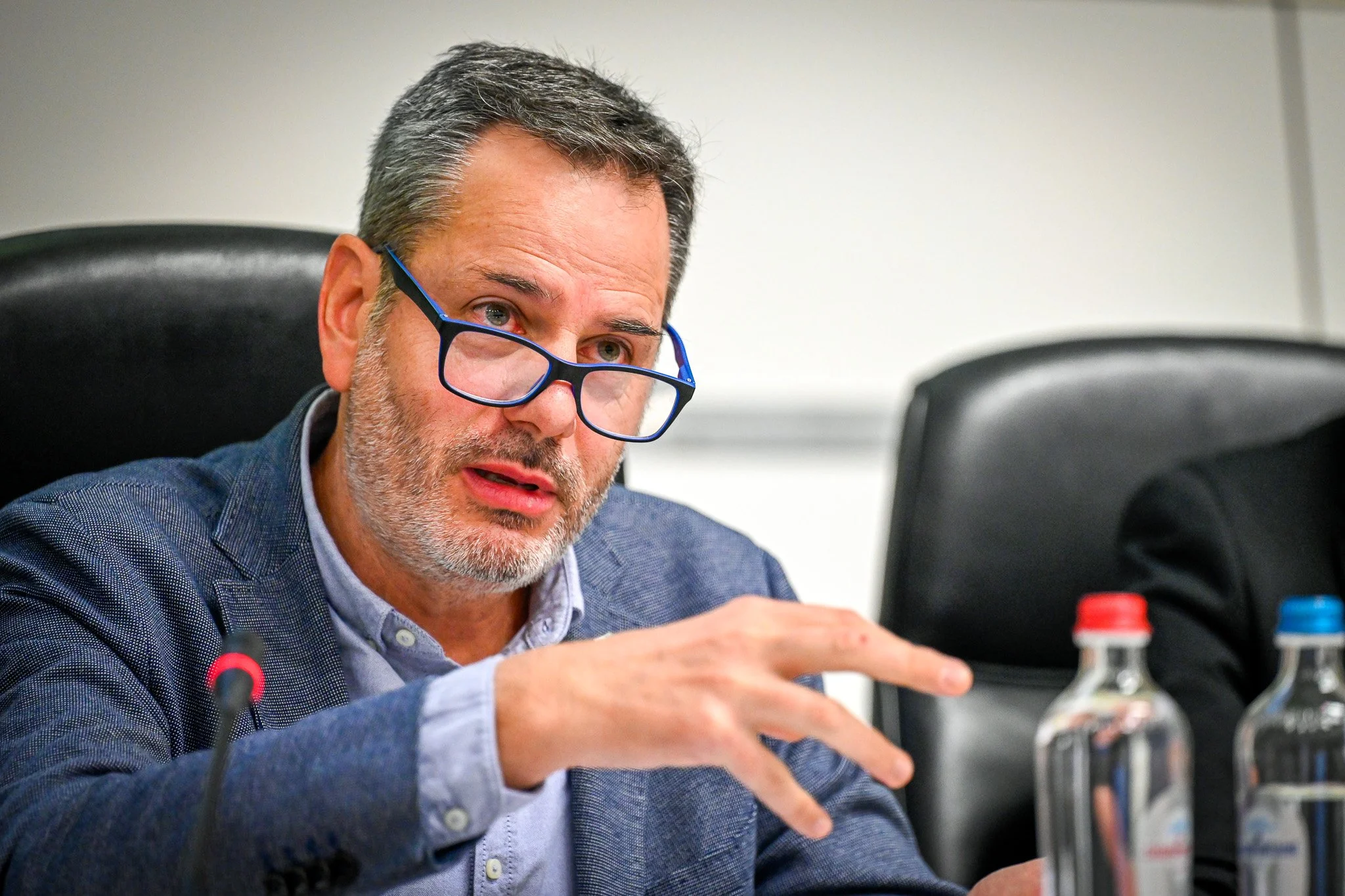 Un homme barbu avec des lunettes bleues parle lors d'une réunion, avec des bouteilles d'eau en plastique sur la table devant lui.
