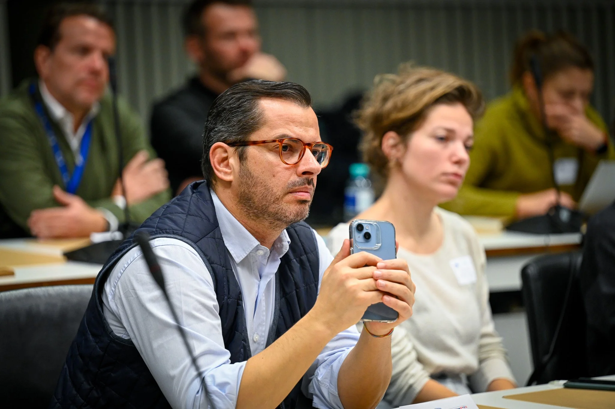 Un homme avec des lunettes tient un smartphone dans une salle de réunion, entouré d'autres personnes assises.