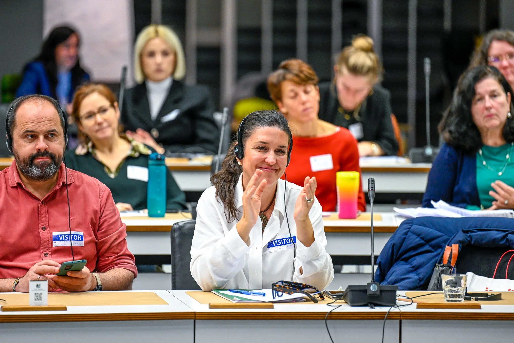 Photo d'une conférence à Bruxelles. Groupe de personnes assises à des tables en conférence, certaines applaudissant, avec des écouteurs et des badges de visiteur.