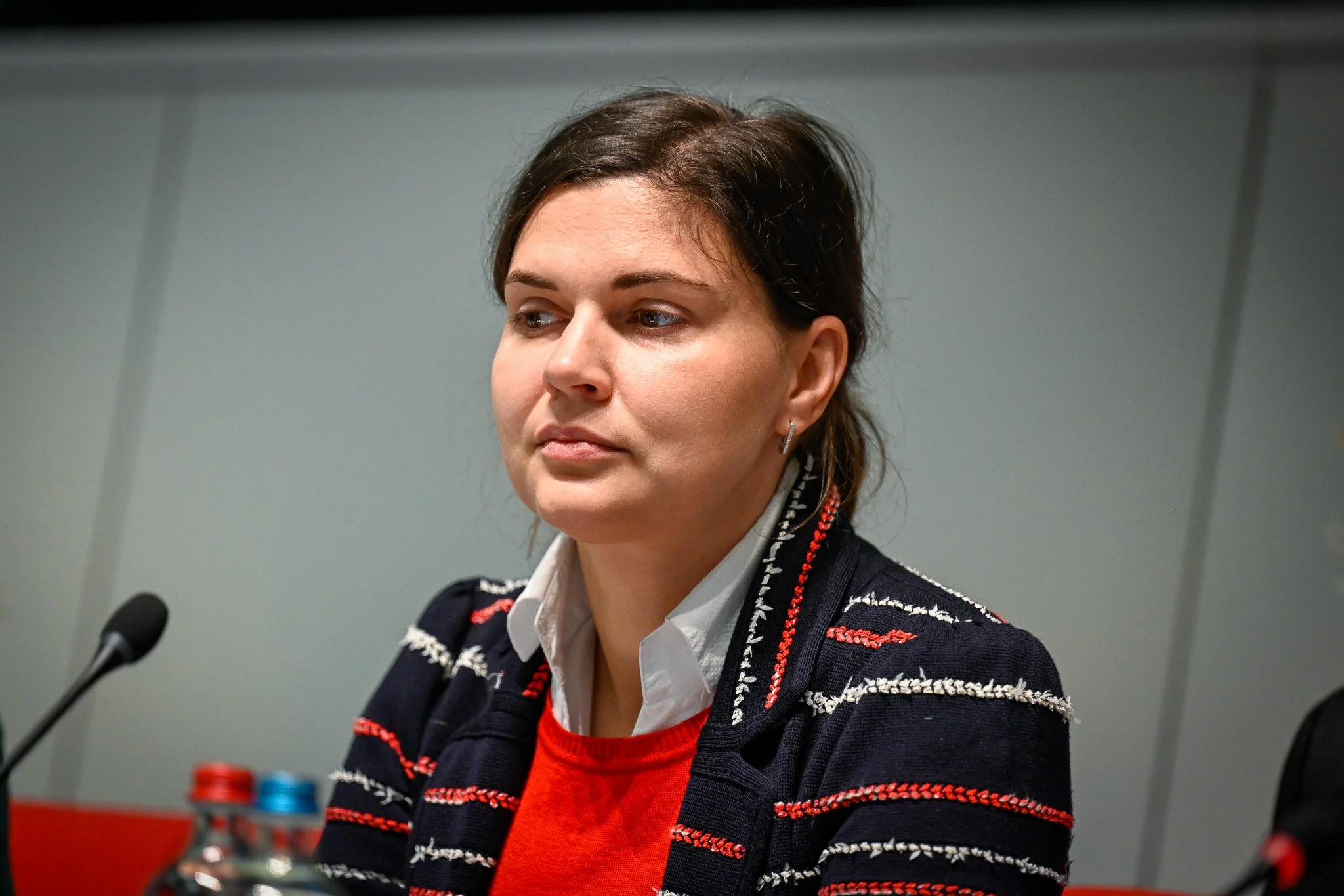 Femme assise devant un microphone, portant une veste rayée et un pull rouge, regardant de côté.