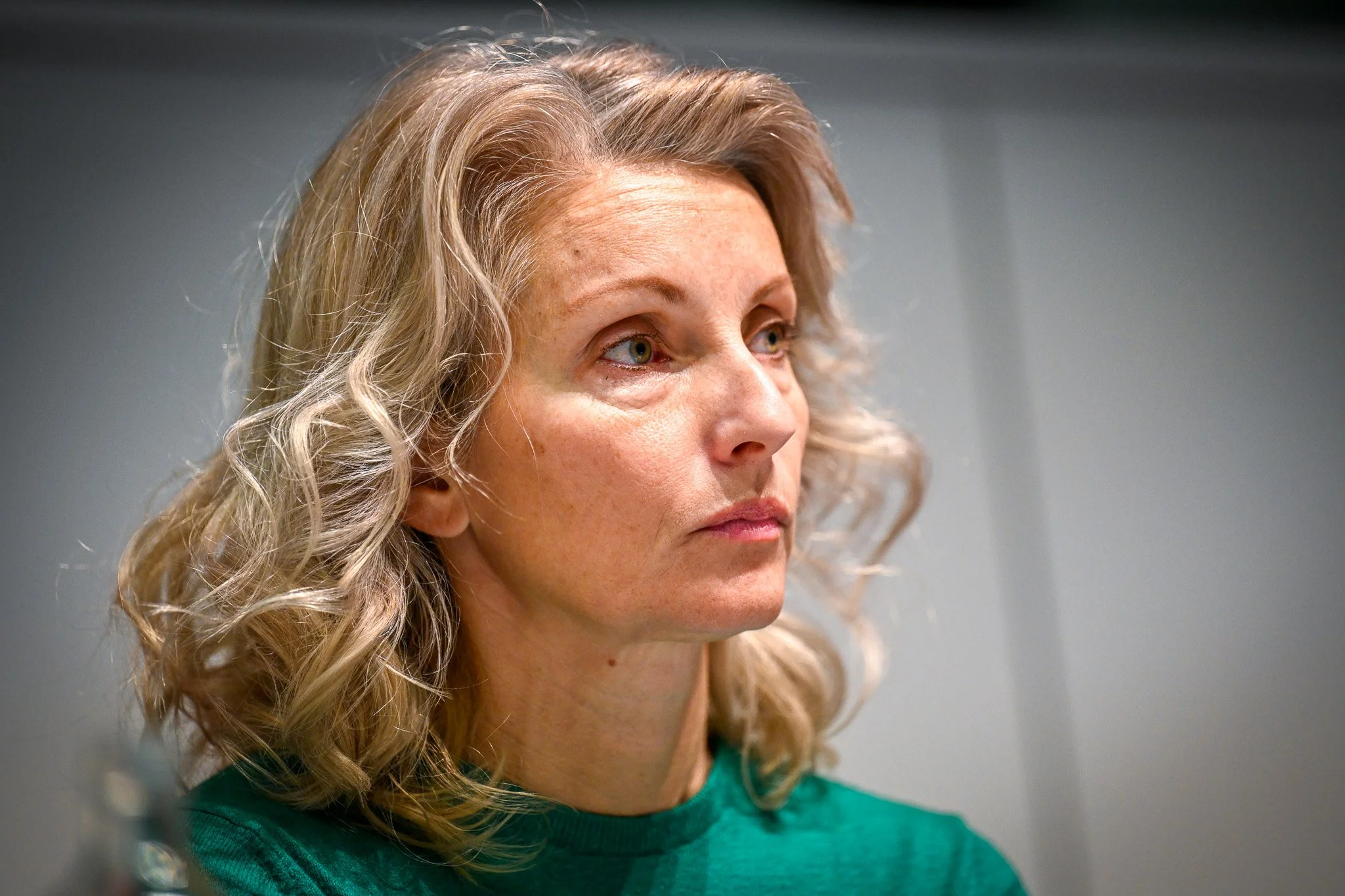 Femme aux cheveux blonds portant un pull vert, regard sérieux.